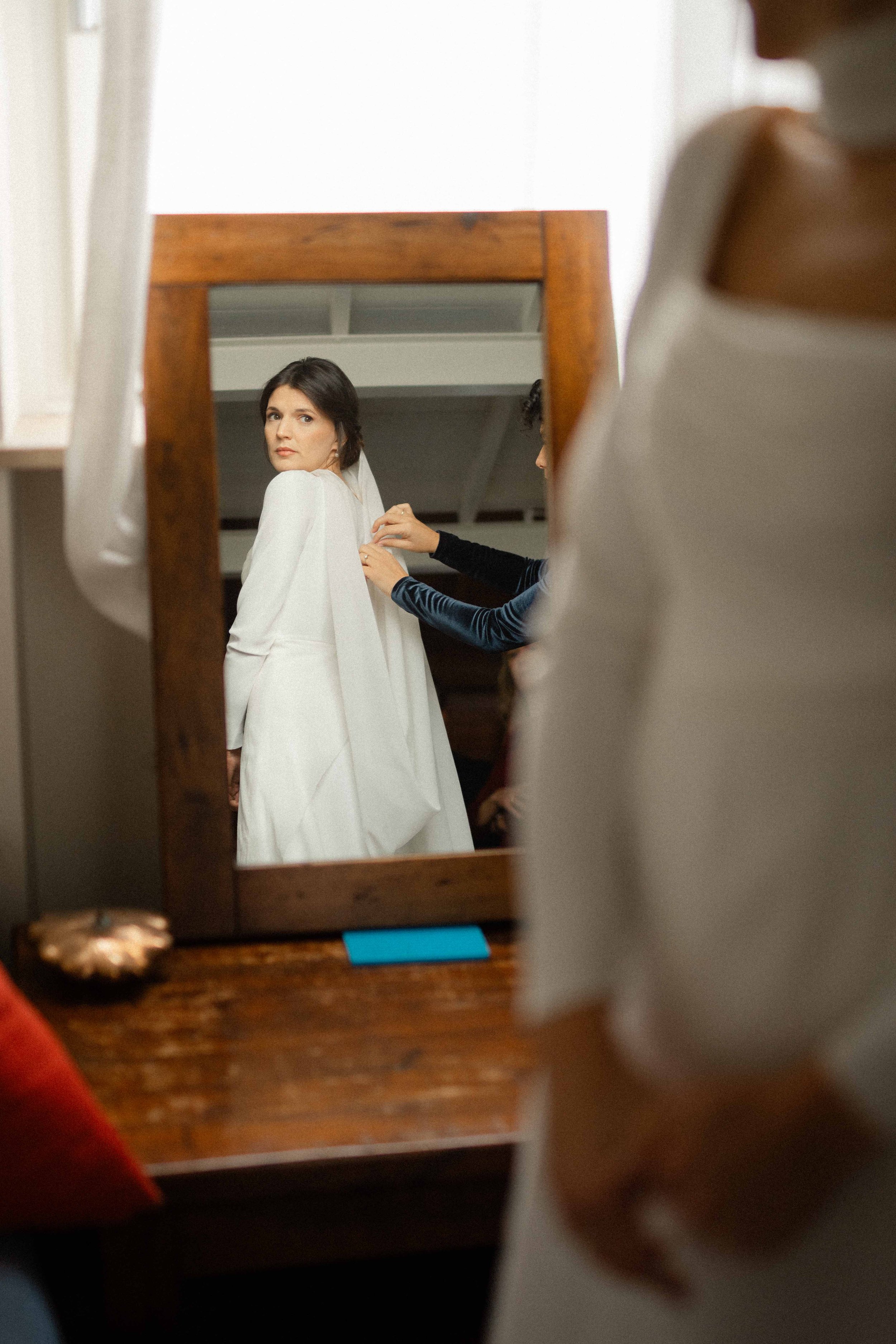 A woman in a white dress is looking at herself in a mirror while someone adjusts her dress.