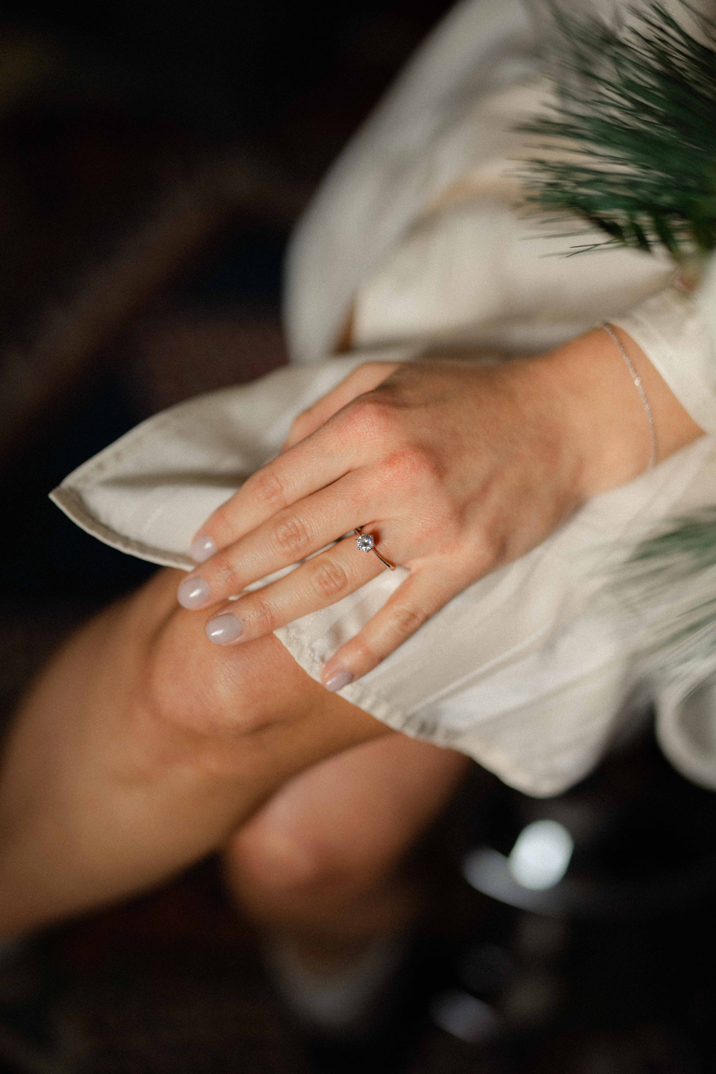 Close-up of a woman's hand with a diamond engagement ring resting on her knee, wearing a cream-colored satin dress with a pearl bracelet, and some greenery in the background.