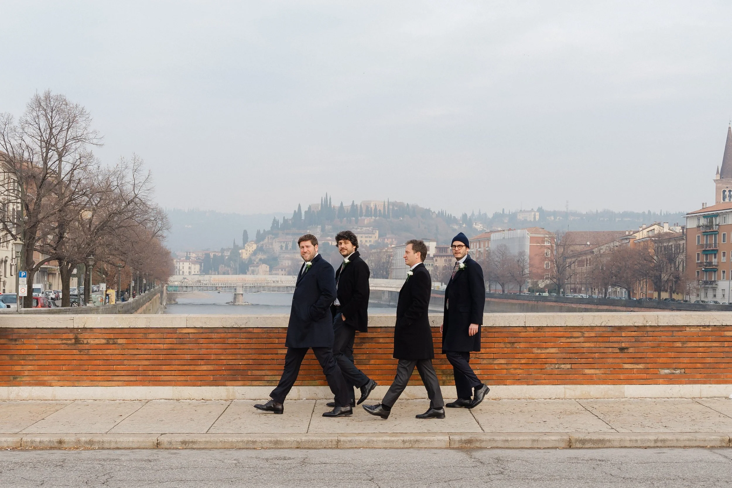 Four men in formal suits walking along a brick wall by a river, with a cityscape and hilly terrain in the background on a cloudy day.