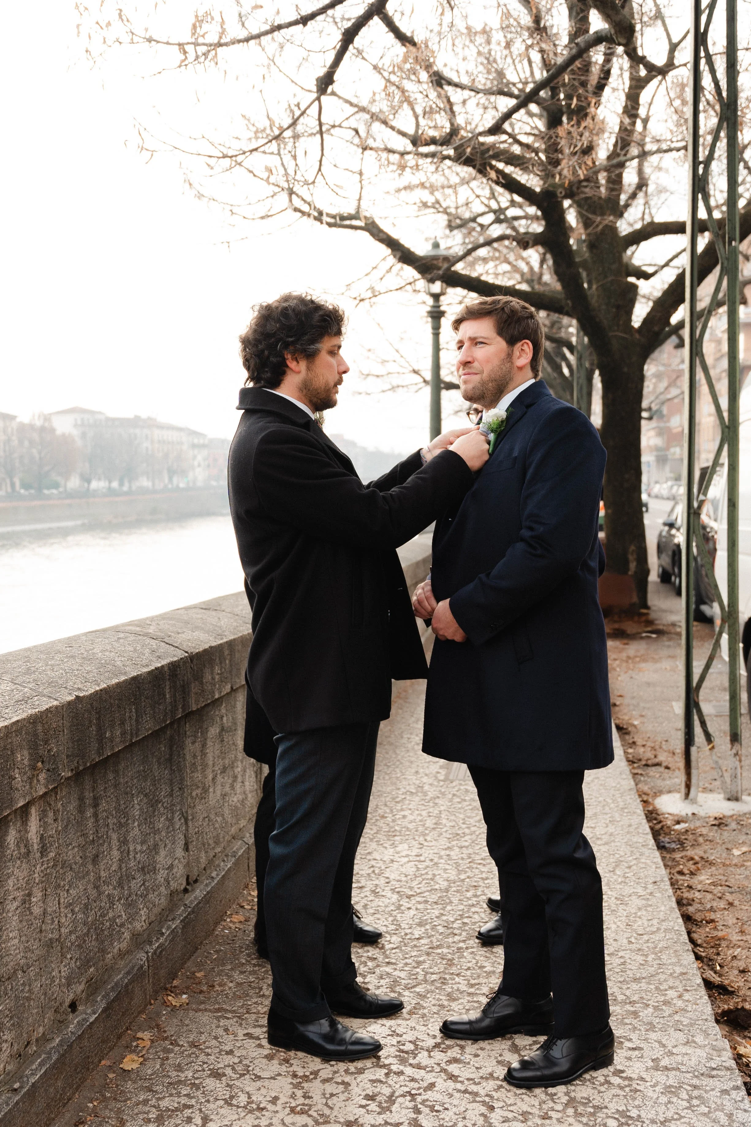Two men dressed in formal attire standing face-to-face on a sidewalk near a river in Verona, North Italy.