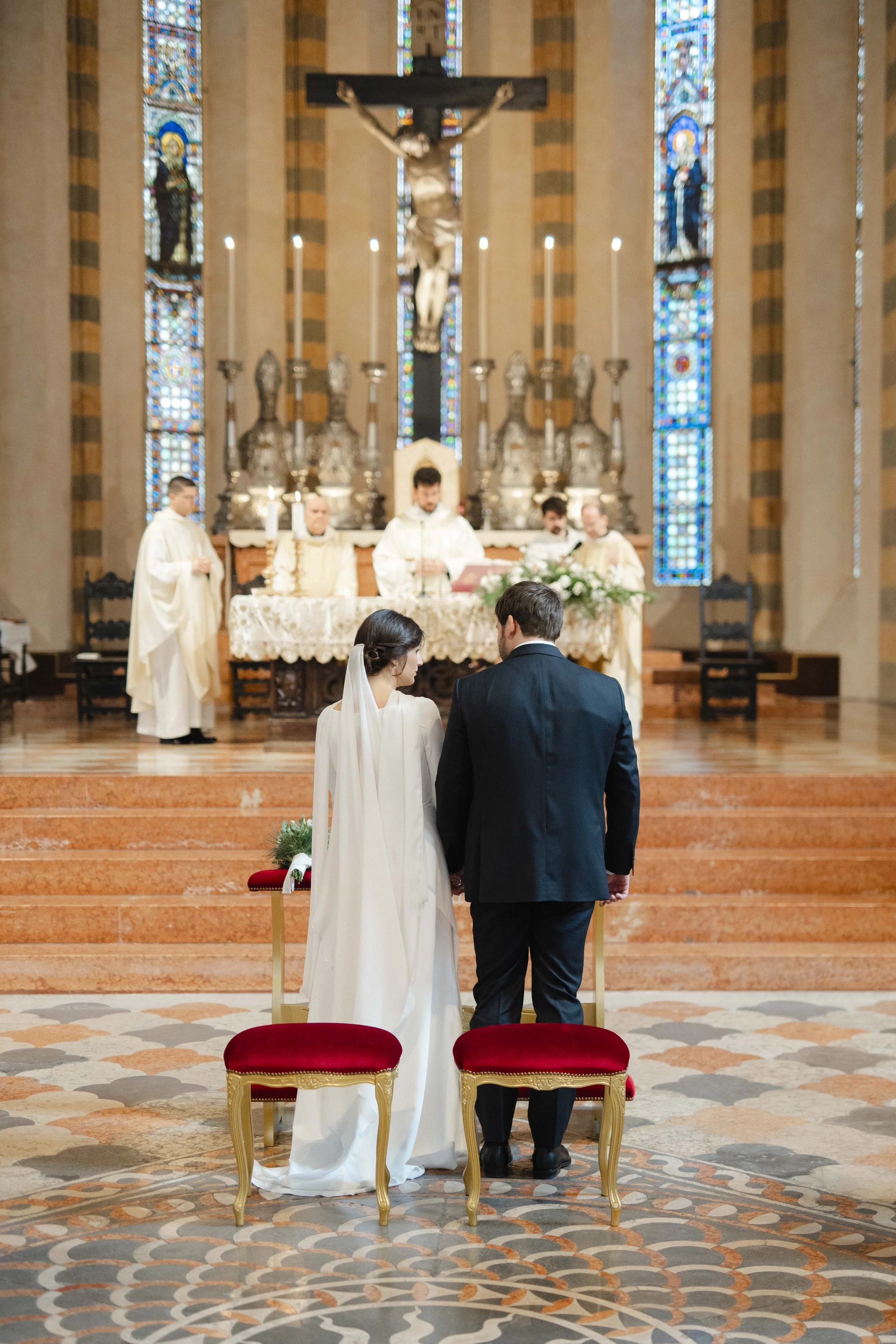 A bride and groom standing before an altar during their wedding ceremony inside a church, with priests and religious figures in the background.