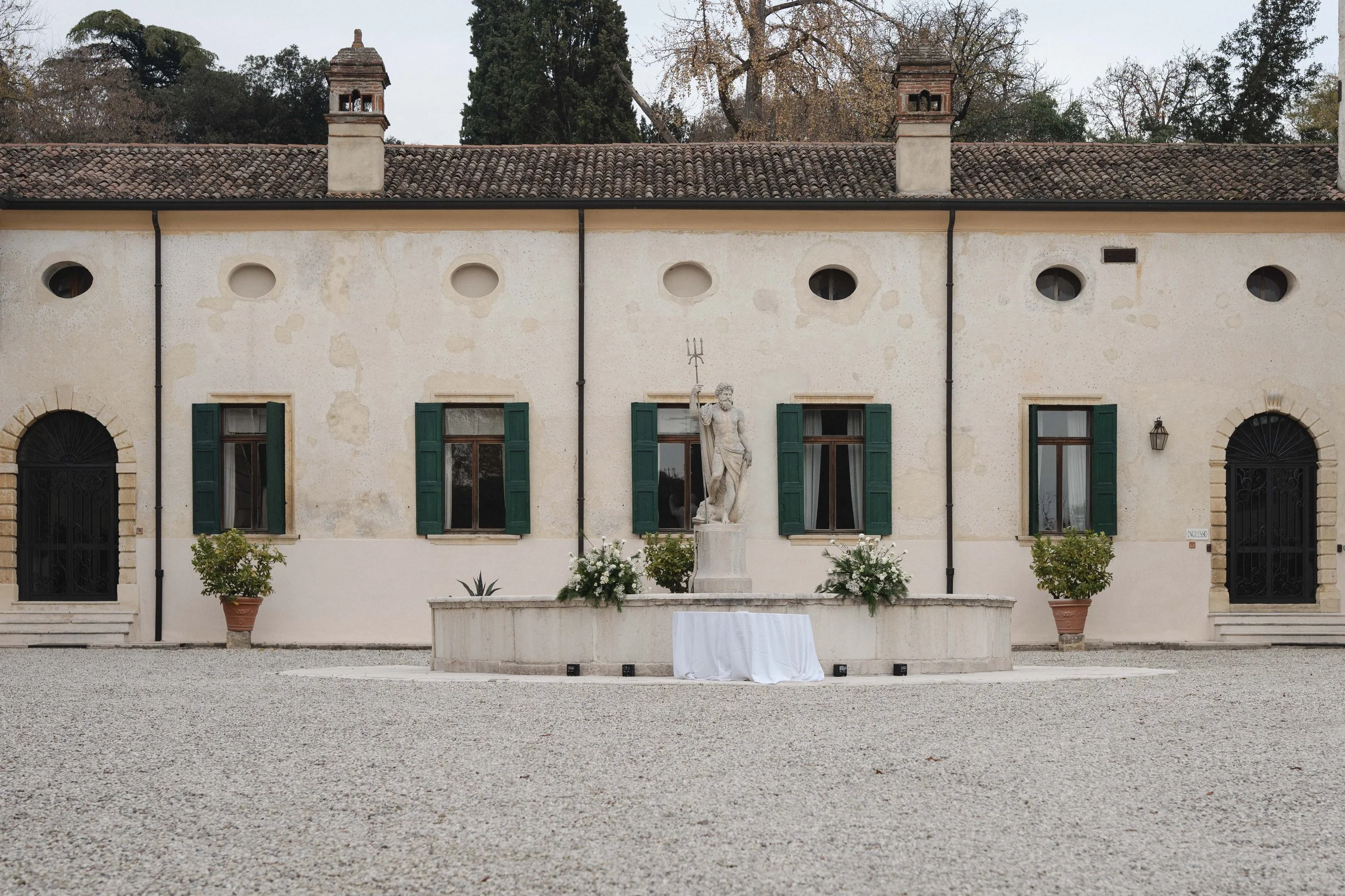 Front view of an old, pale yellow building with green shutters, arched doorways, and a statue of a man holding a trident in the center. The building has a red tile roof with two small chimneys and is surrounded by potted plants and flowers, with trees in the background.