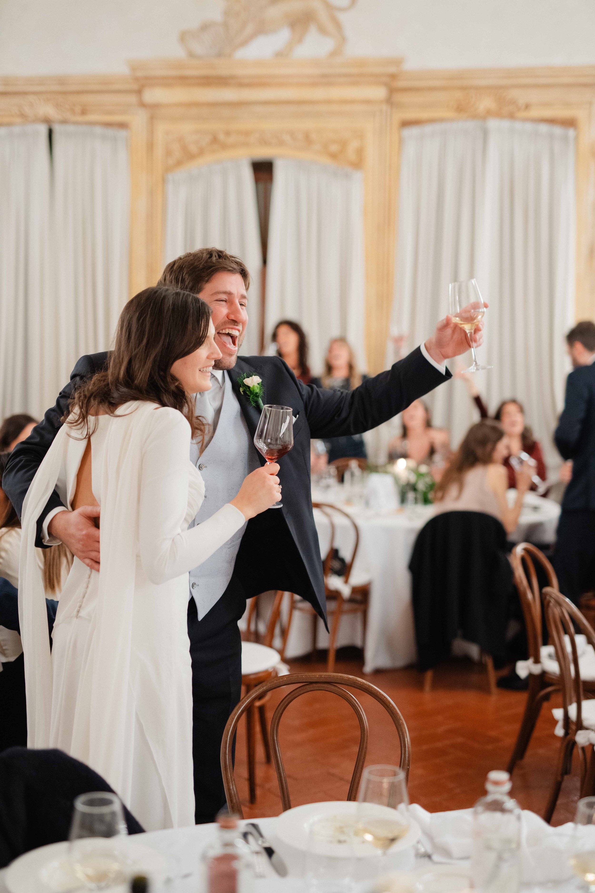 A man and woman at a wedding reception, smiling and raising a glass in celebration. People are seated at tables in the background.