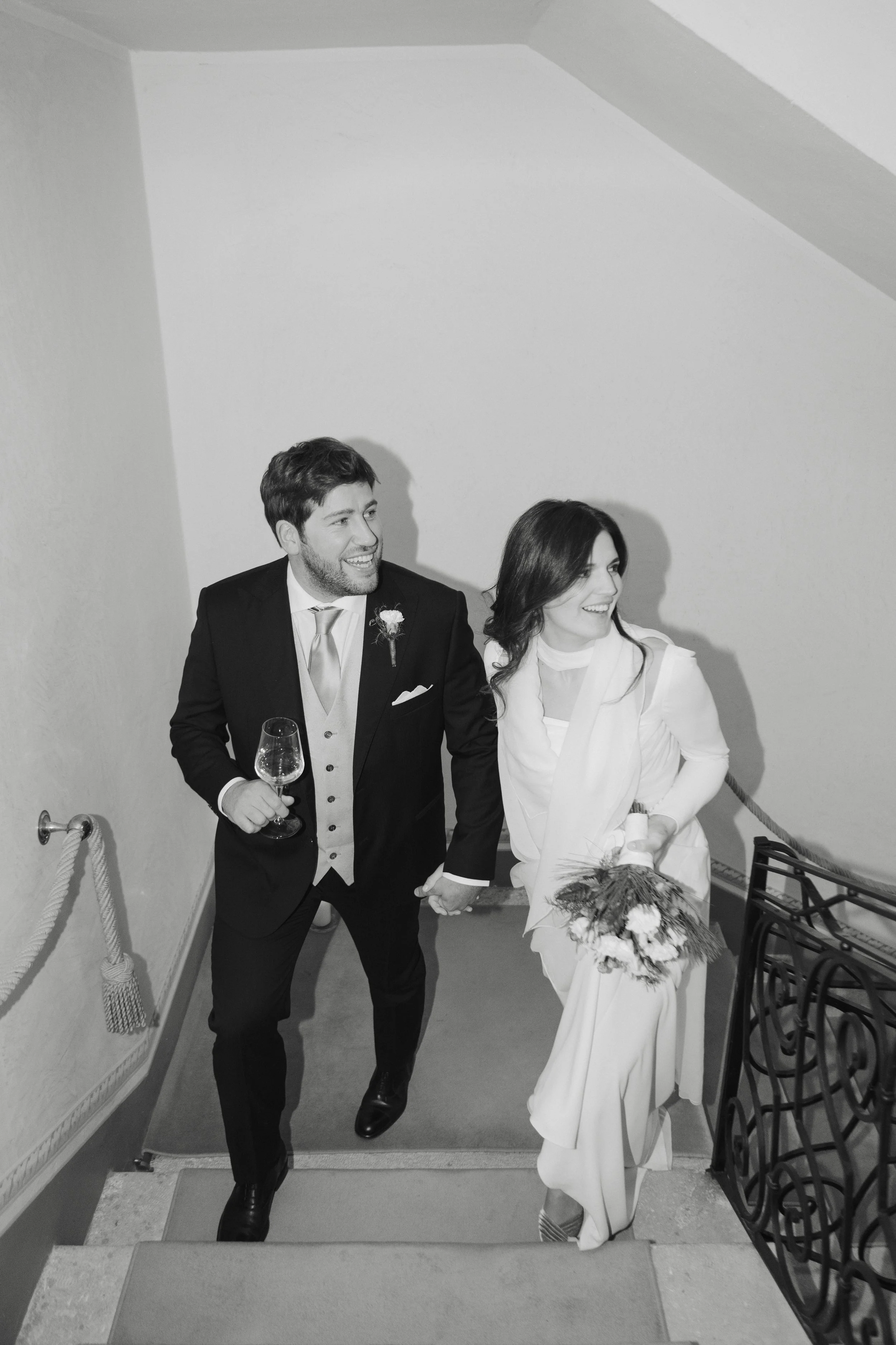 A happy man and woman holding hands while walking up the stairs after a wedding, with the man holding a wine glass and both dressed in formal wedding attire.