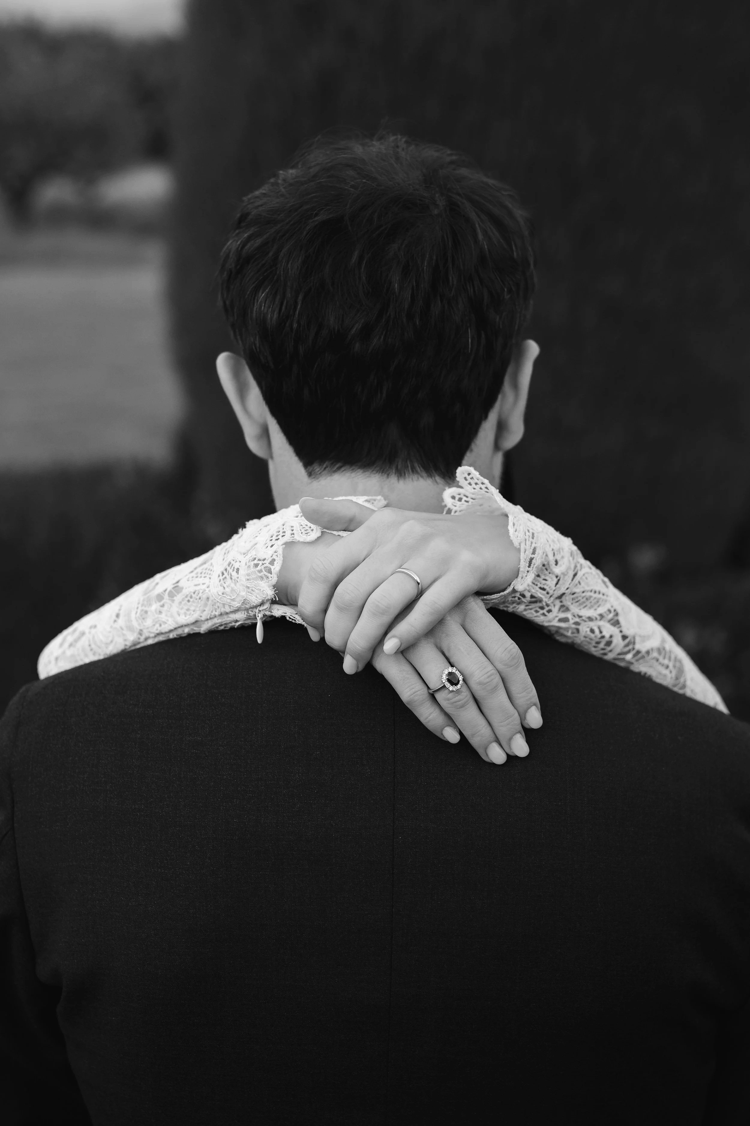 Back of a person's head with short dark hair, wearing a dark blazer, and a hand with rings resting on their shoulder, with lace garment and rings visible, in black and white.