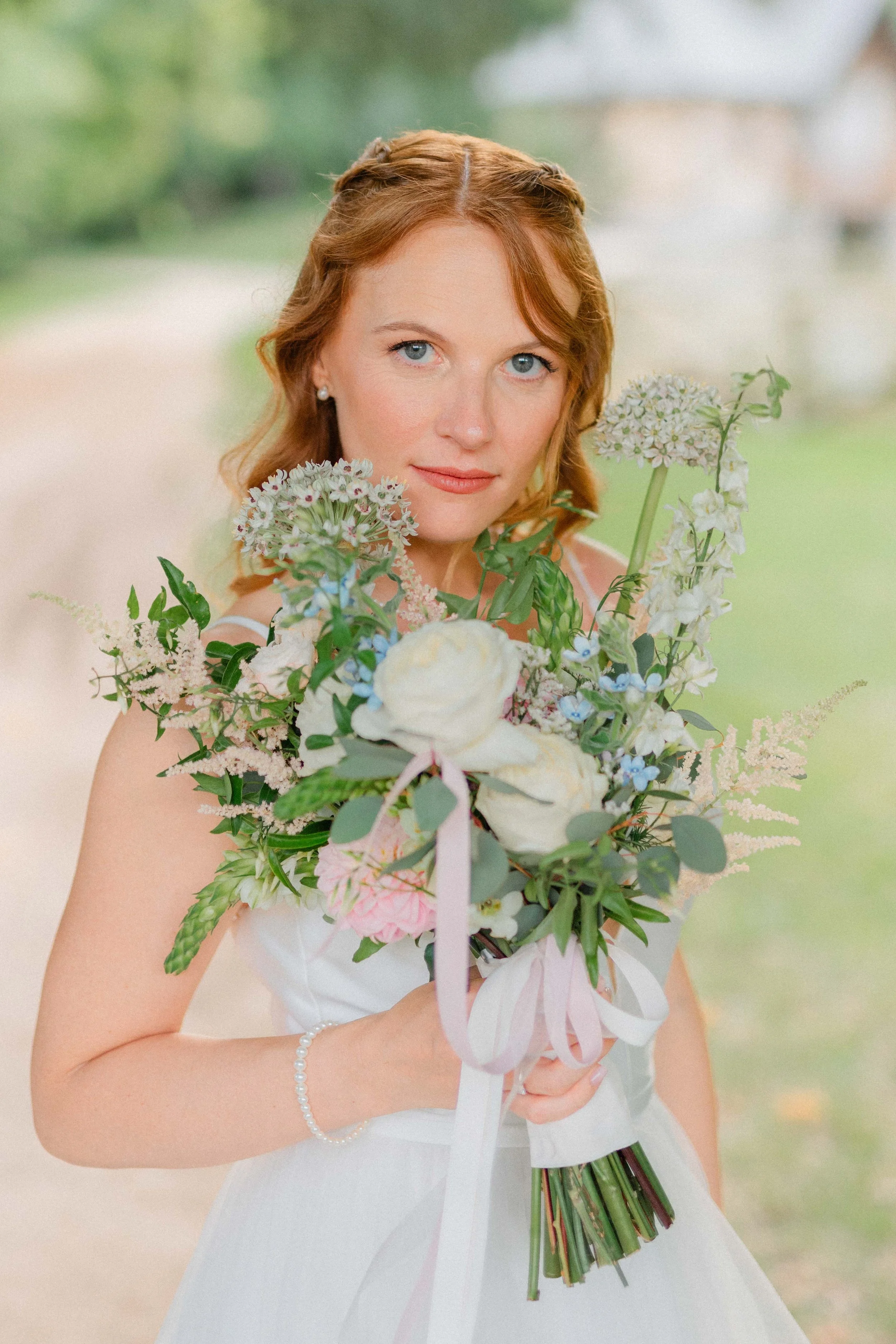 A woman with red hair and blue eyes holding a large bouquet of flowers, wearing a white dress and pearl bracelet outdoors.