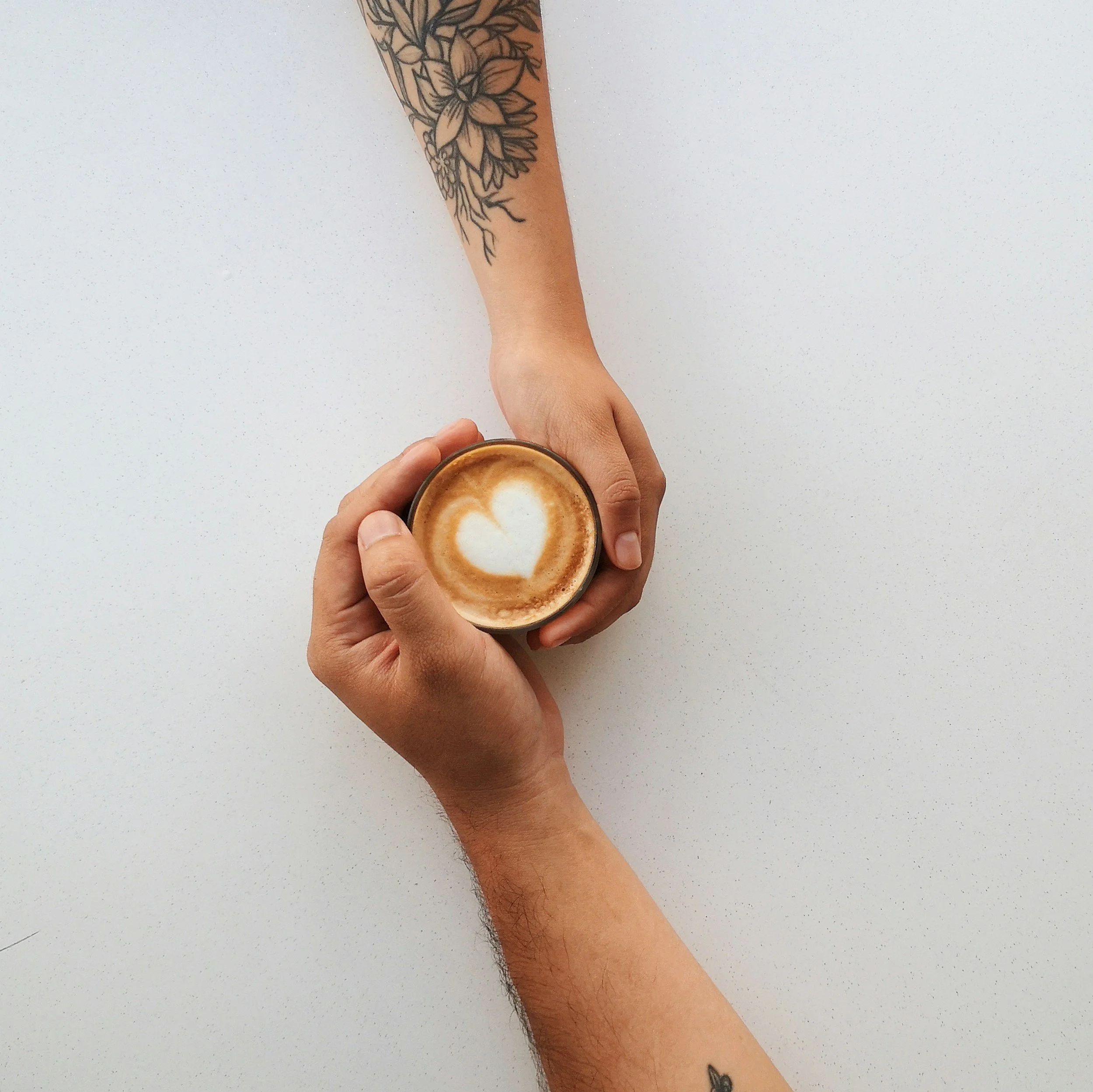 A person's arm with a tattoo of flowers, holding a cup of coffee on a white speckled surface.