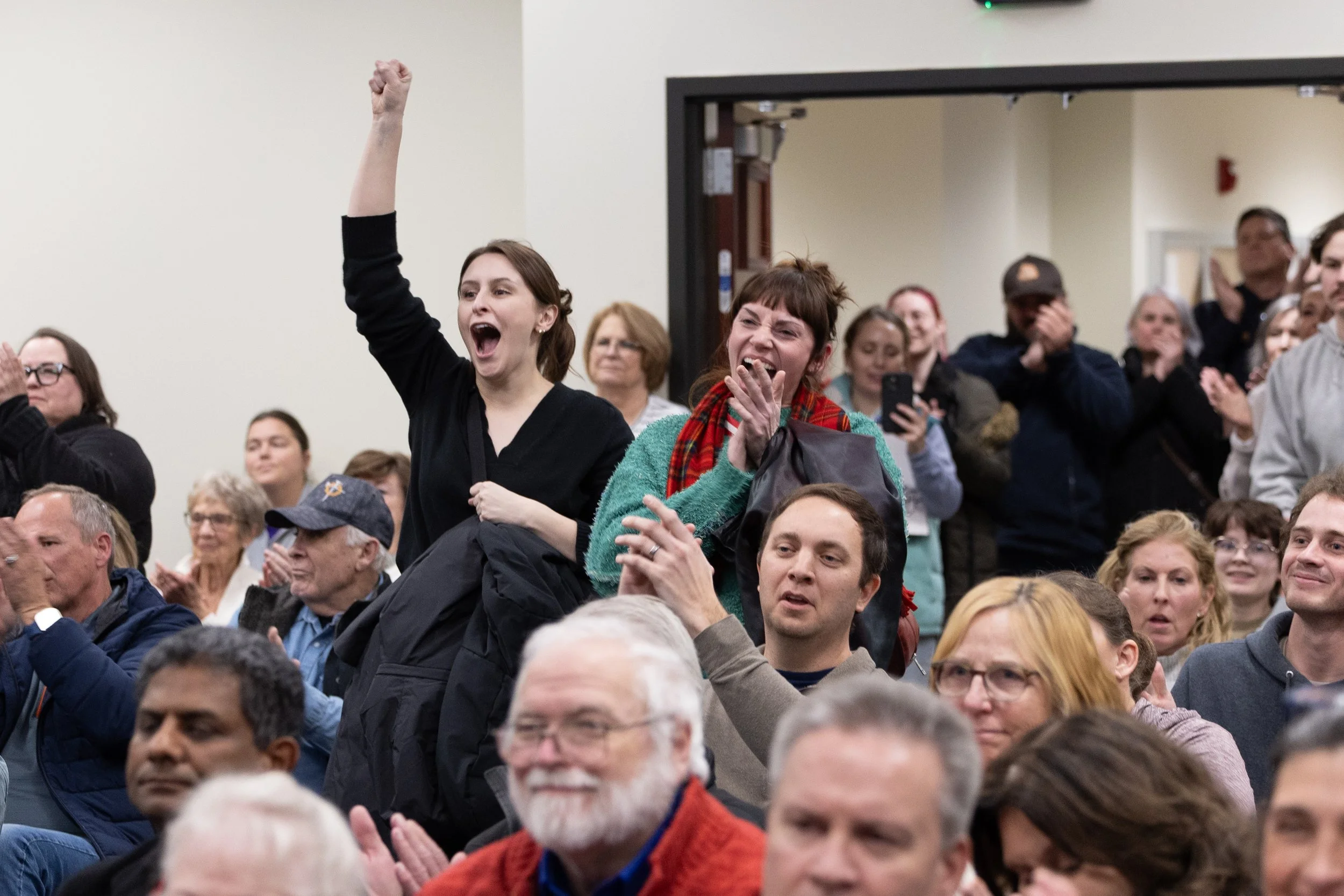 A woman with brown hair and a black top is cheering with her mouth open and her right arm raised in a fist. She is surrounded by people, some of whom are clapping or smiling, in a city hall conference room.