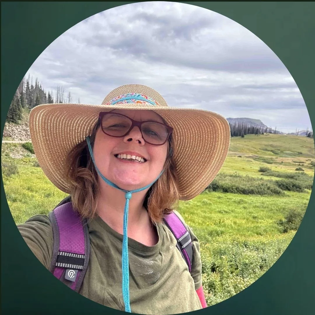 Headshot of smiling educator in front of an open field wearing a floppy hat.