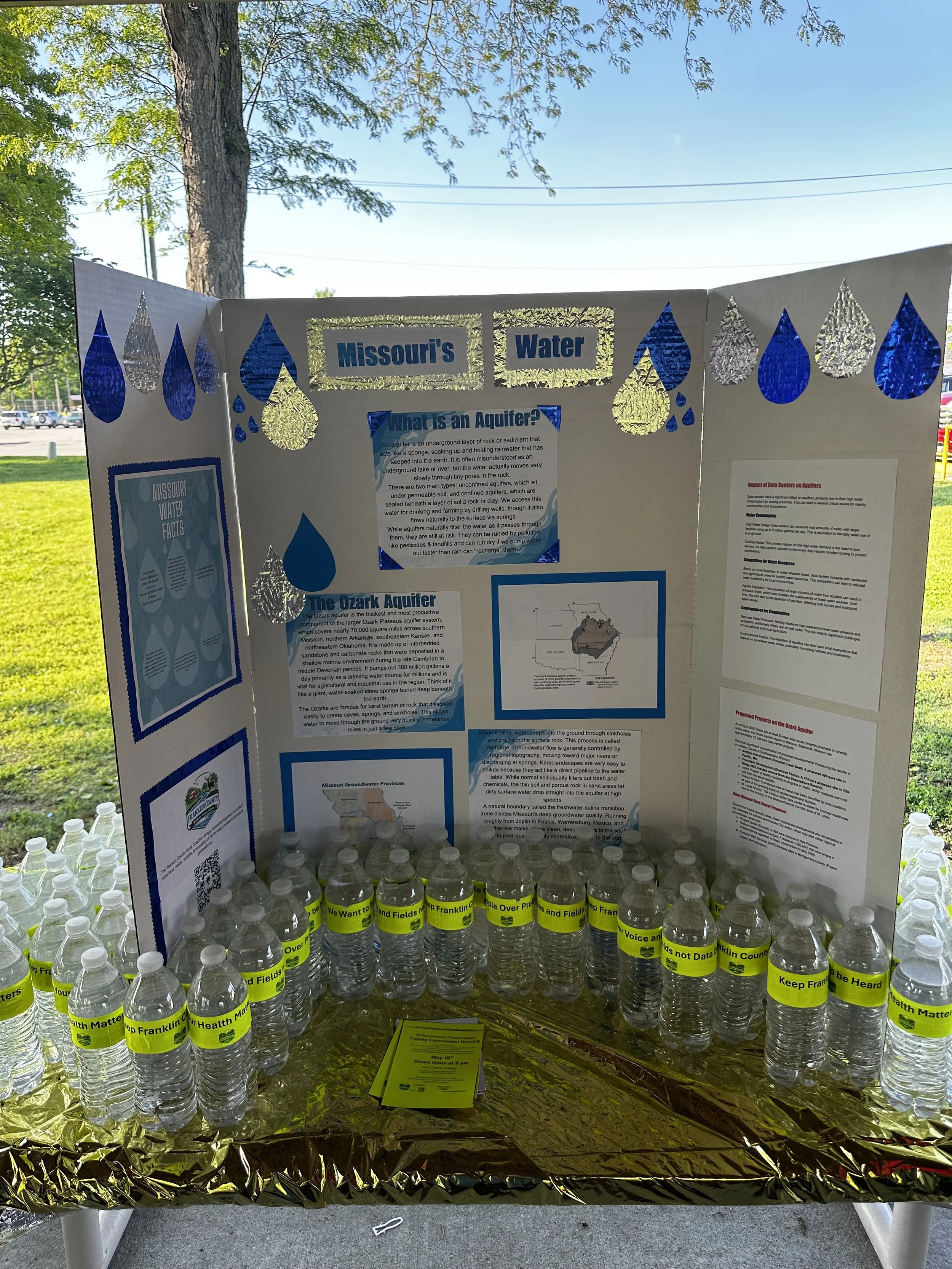 An informational display about Missouri water aquifers at an outdoor event, with bottles of water and brochures on a picnic table under a park pavilion.