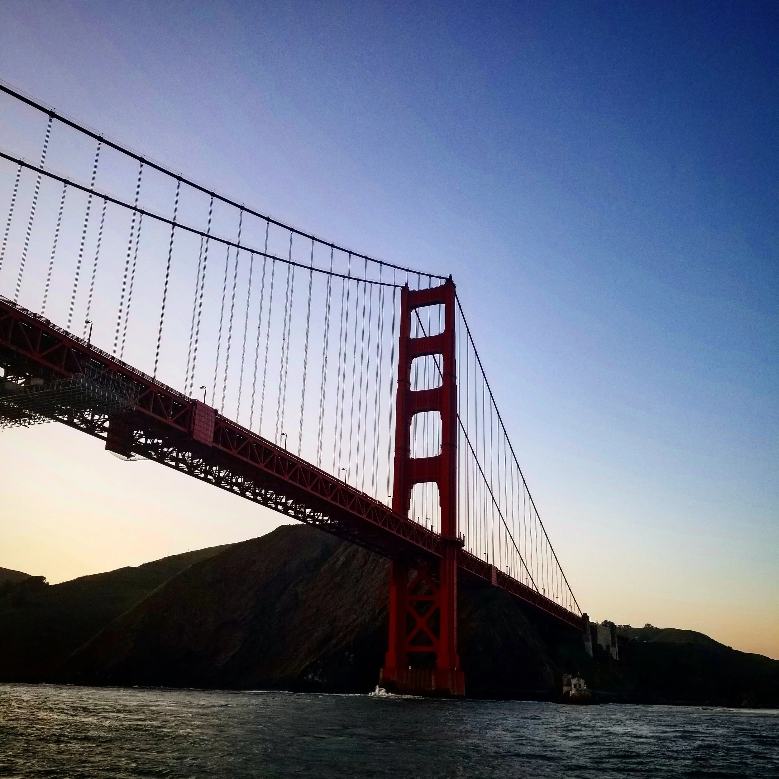 A photo of the Golden Gate Bridge against a darkening sky, viewed from the water below with hills in the background.