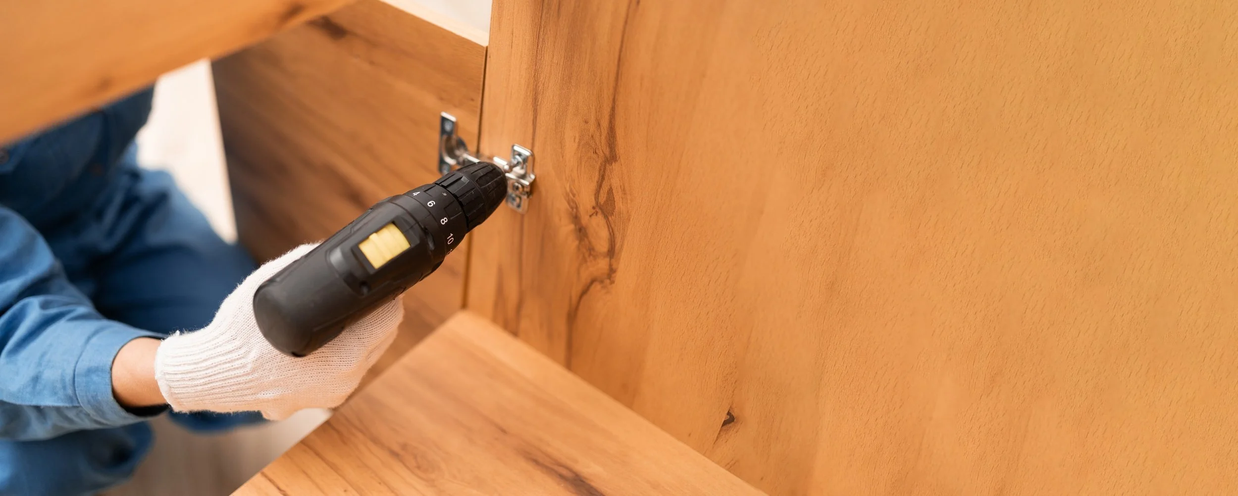 A person wearing a white glove and blue shirt using a cordless drill to secure a hinge onto a wooden cabinet door.