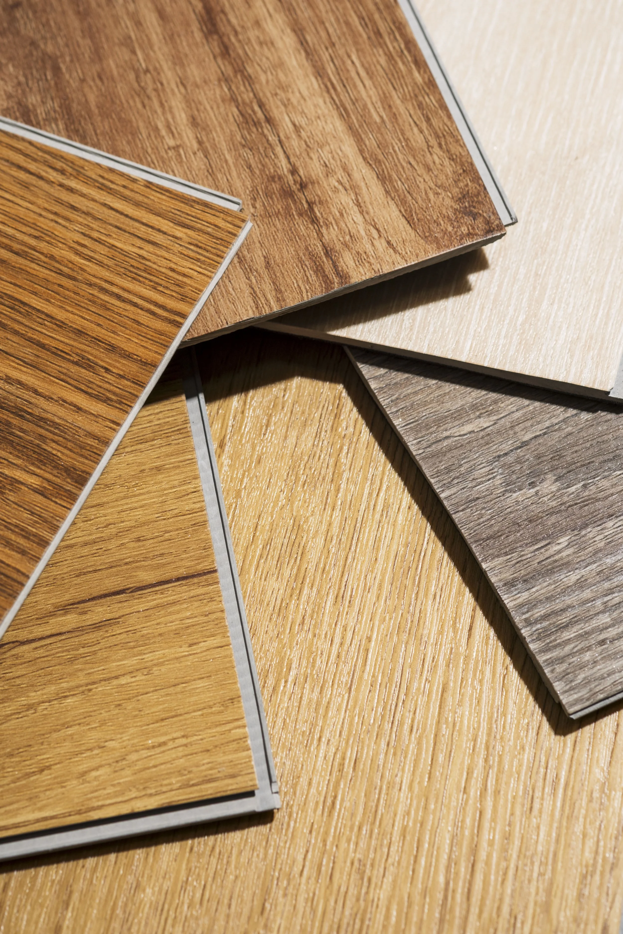 Multiple wood laminate flooring samples in various shades and grains are spread out on a wooden surface.