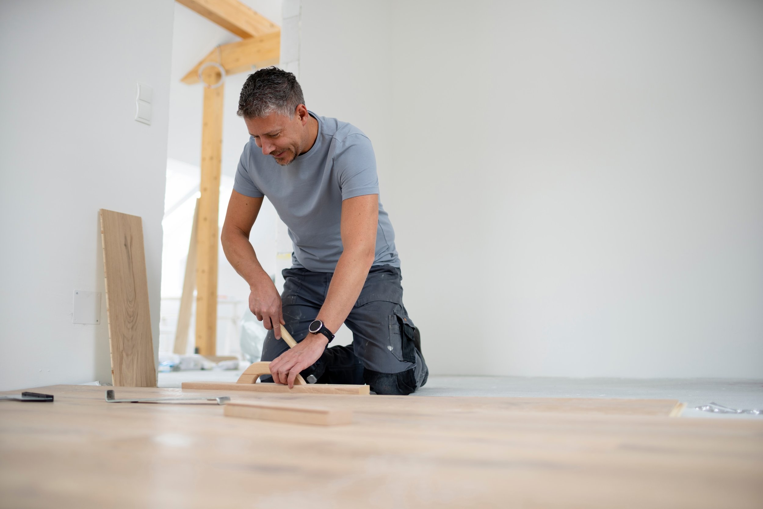 A man working on a woodworking project indoors, using a saw on a piece of wood, with tools and wood pieces on the floor.