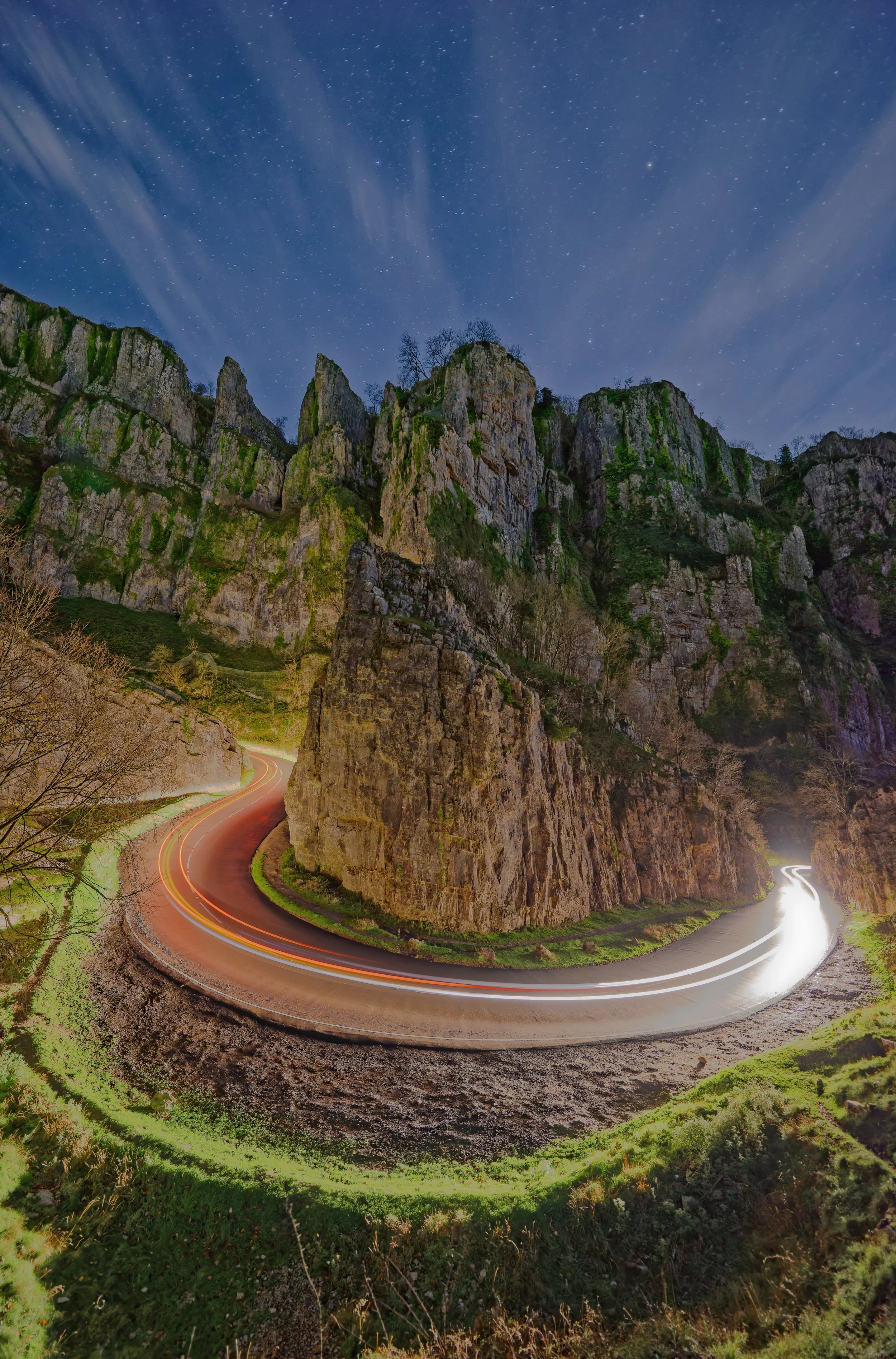 Red and white vehicle light trails curve around Cheddar Gorge beneath rugged cliffs and a starry blue night sky, Somerset