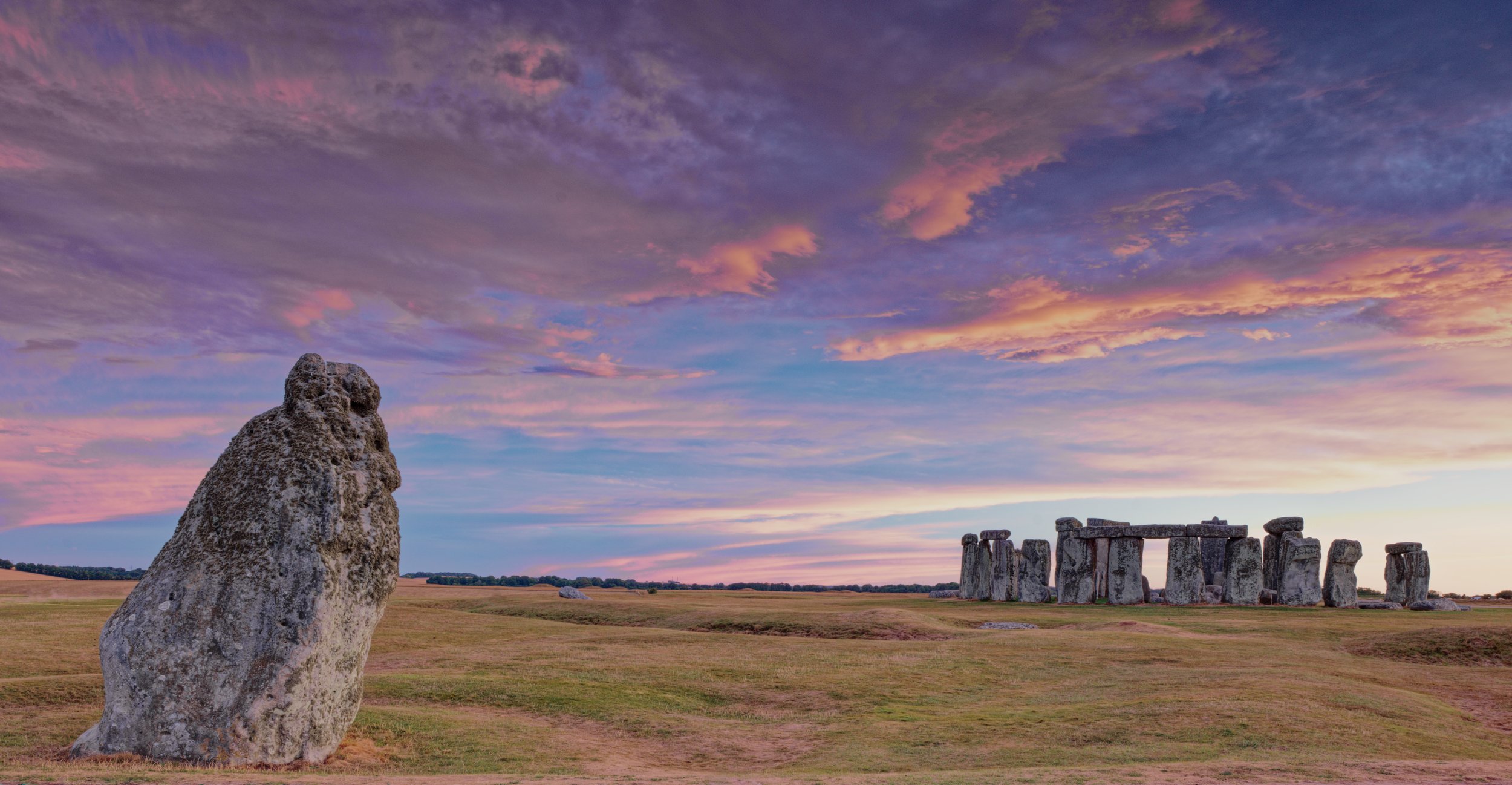 The Heel Stone in the foreground with Stonehenge beneath sweeping pink and violet clouds in Wiltshire