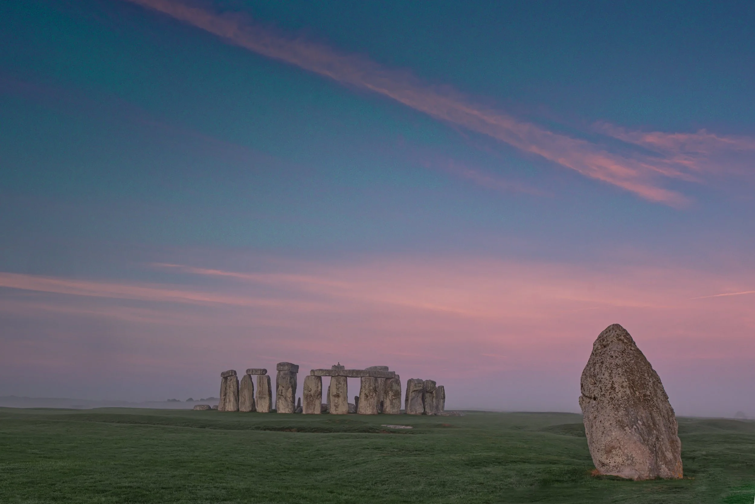 Stonehenge and the Heel Stone on green grass beneath blue skies and rose-pink clouds in Wiltshire