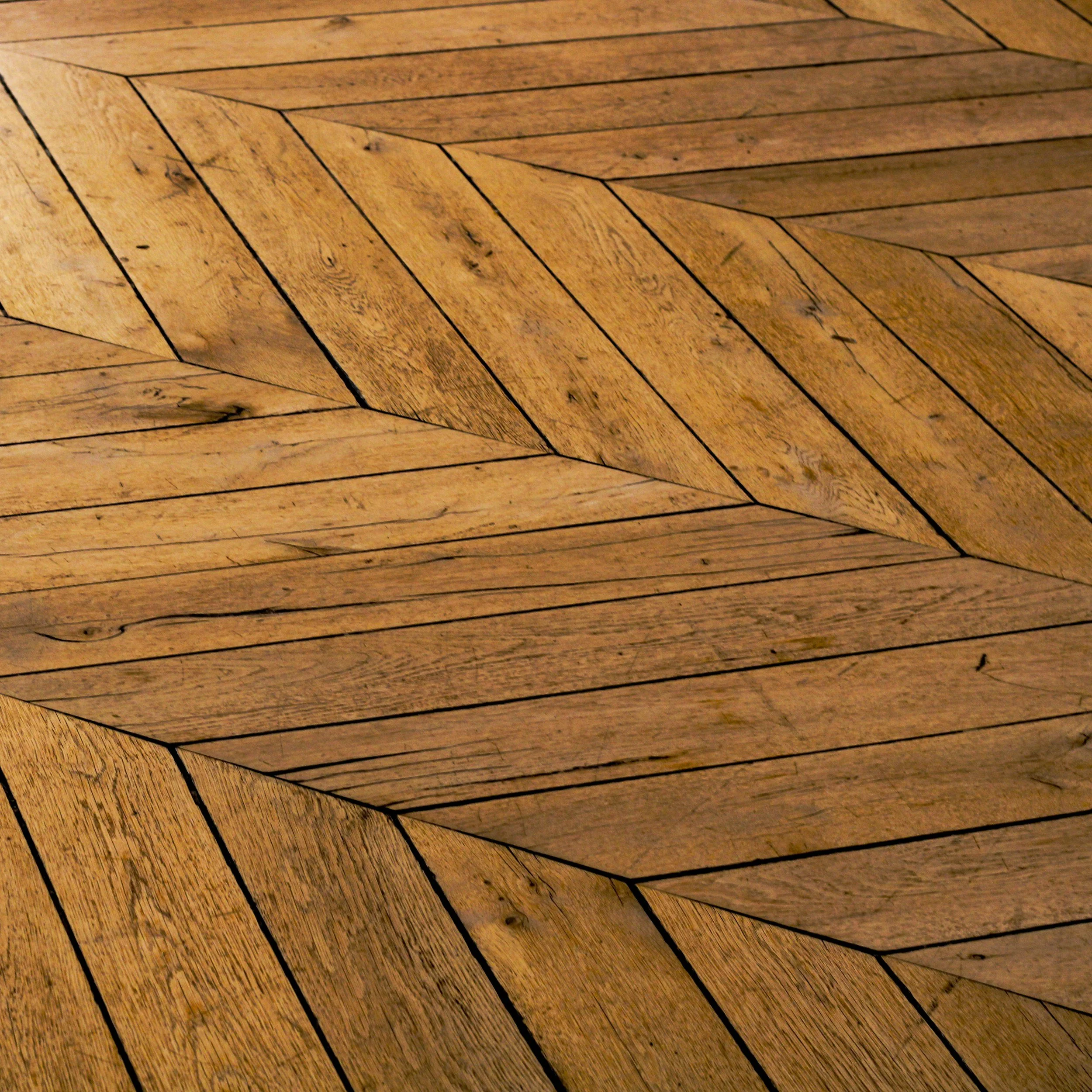 Close-up photo of a wooden parquet flooring with a herringbone pattern, showing the natural wood grain and color variations.