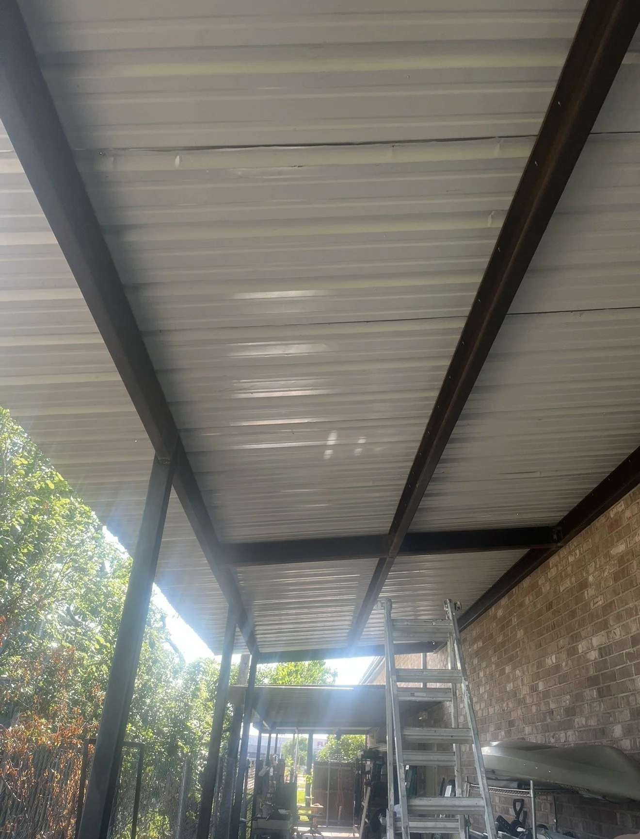 Metal roofed patio extension under construction with scaffolding and ladder, brick wall on the side, and trees in the background.