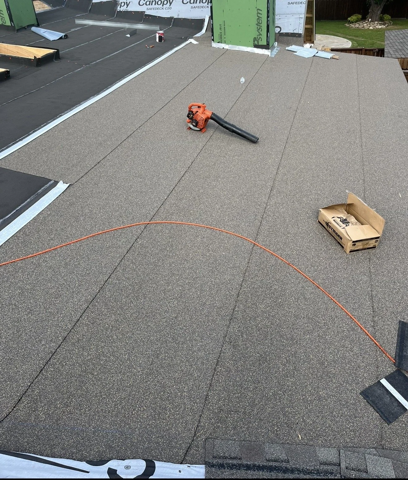Roof being installed with new shingles, construction tools, a box, and a ladder visible in the background.