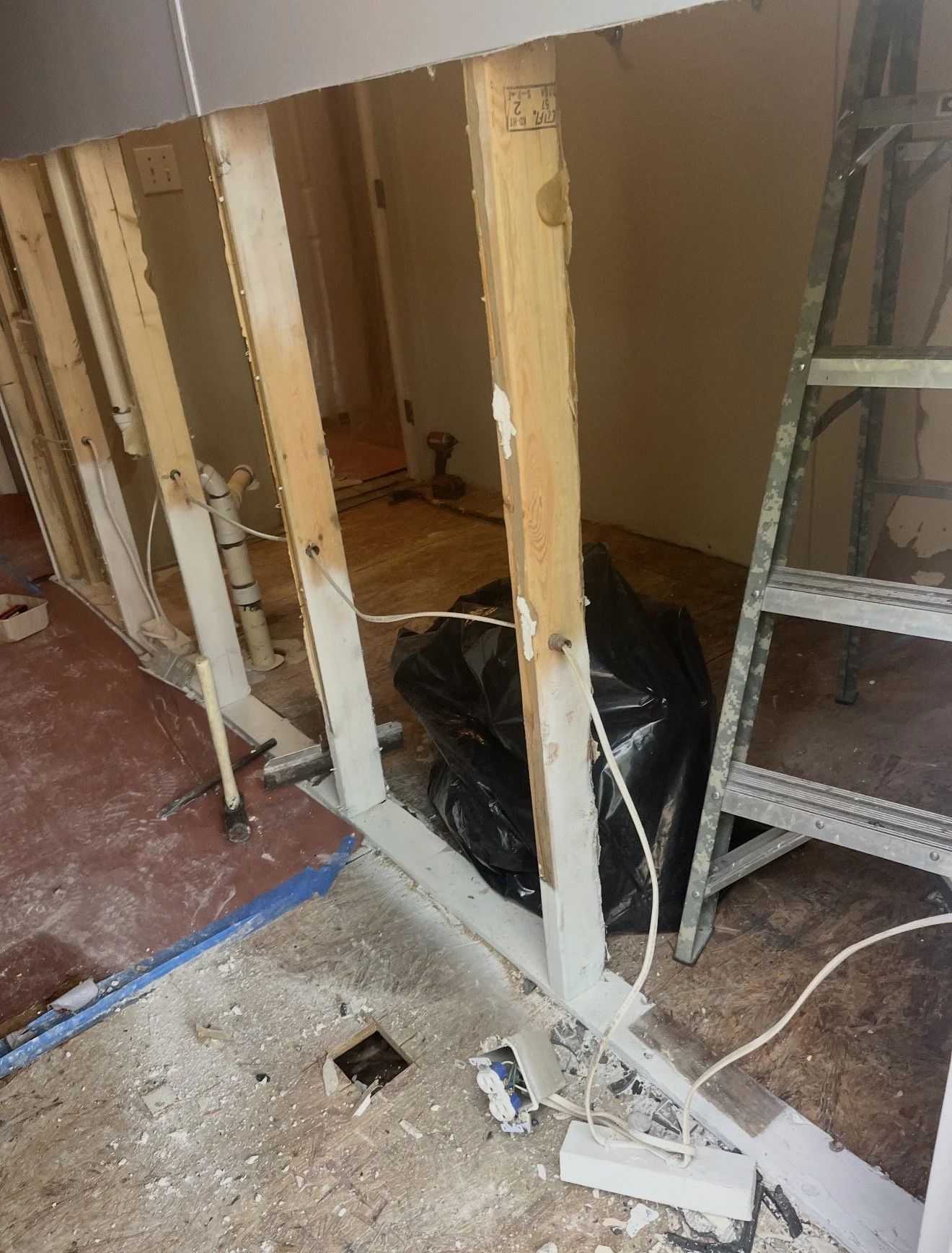 Partially demolished interior wall with exposed studs, pipes, electrical wiring, and tools, indicating ongoing renovation work.