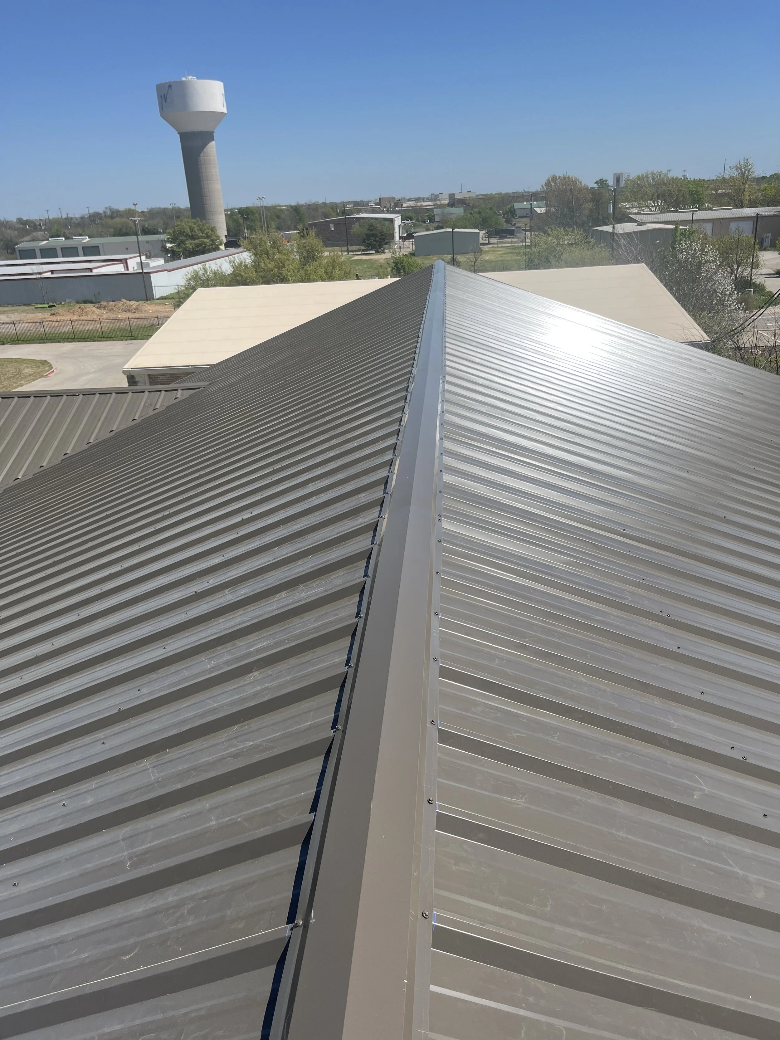 View from a building’s roof showing a metal rooftop with a clear blue sky, trees, and a water tower in the background.
