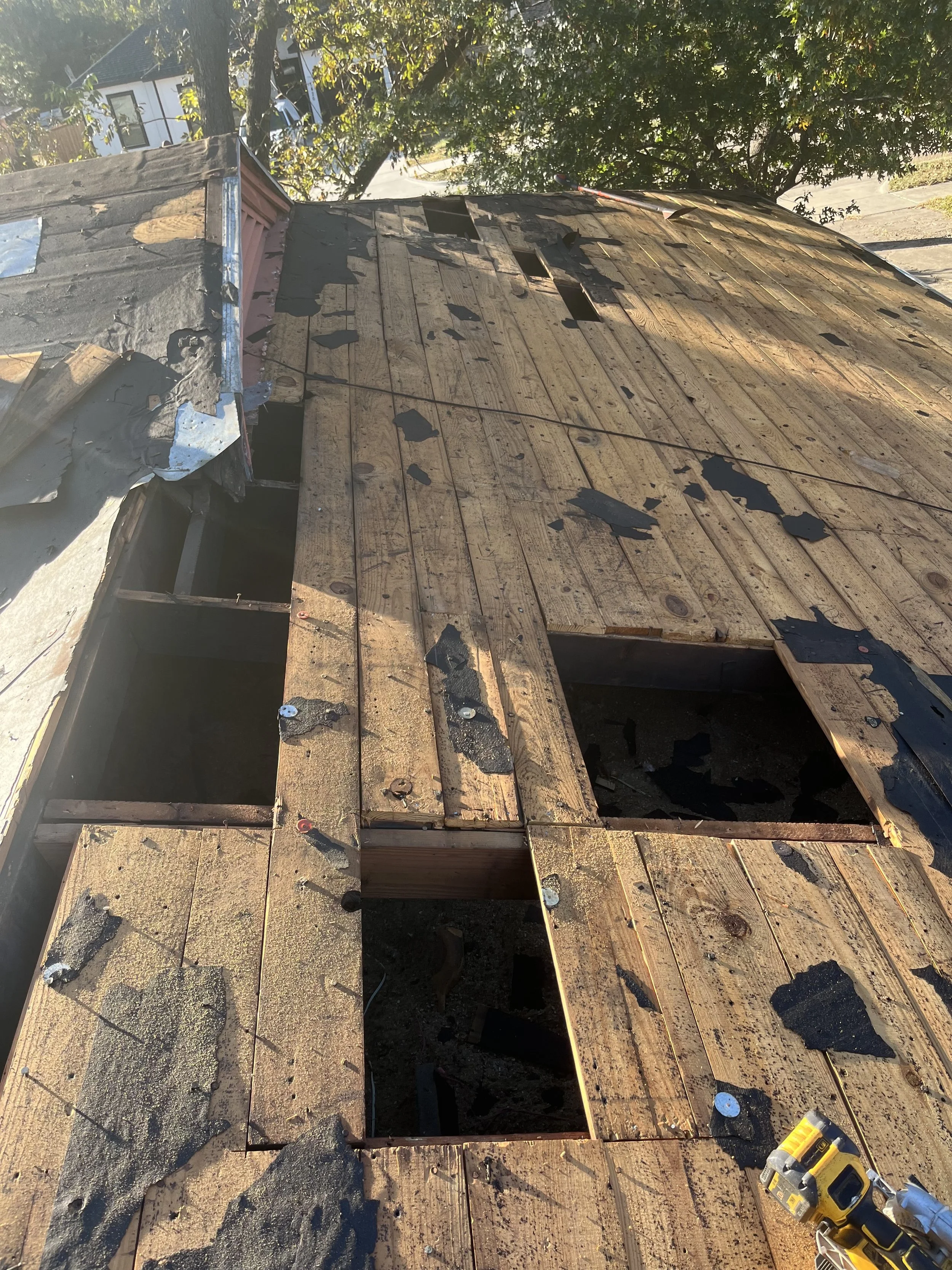 A wooden roof is being repaired with some shingles removed. Tools are visible on the right side of the image, including a yellow drill. The area shows exposed wood and missing shingles, with some shingles in a pile on the left side.
