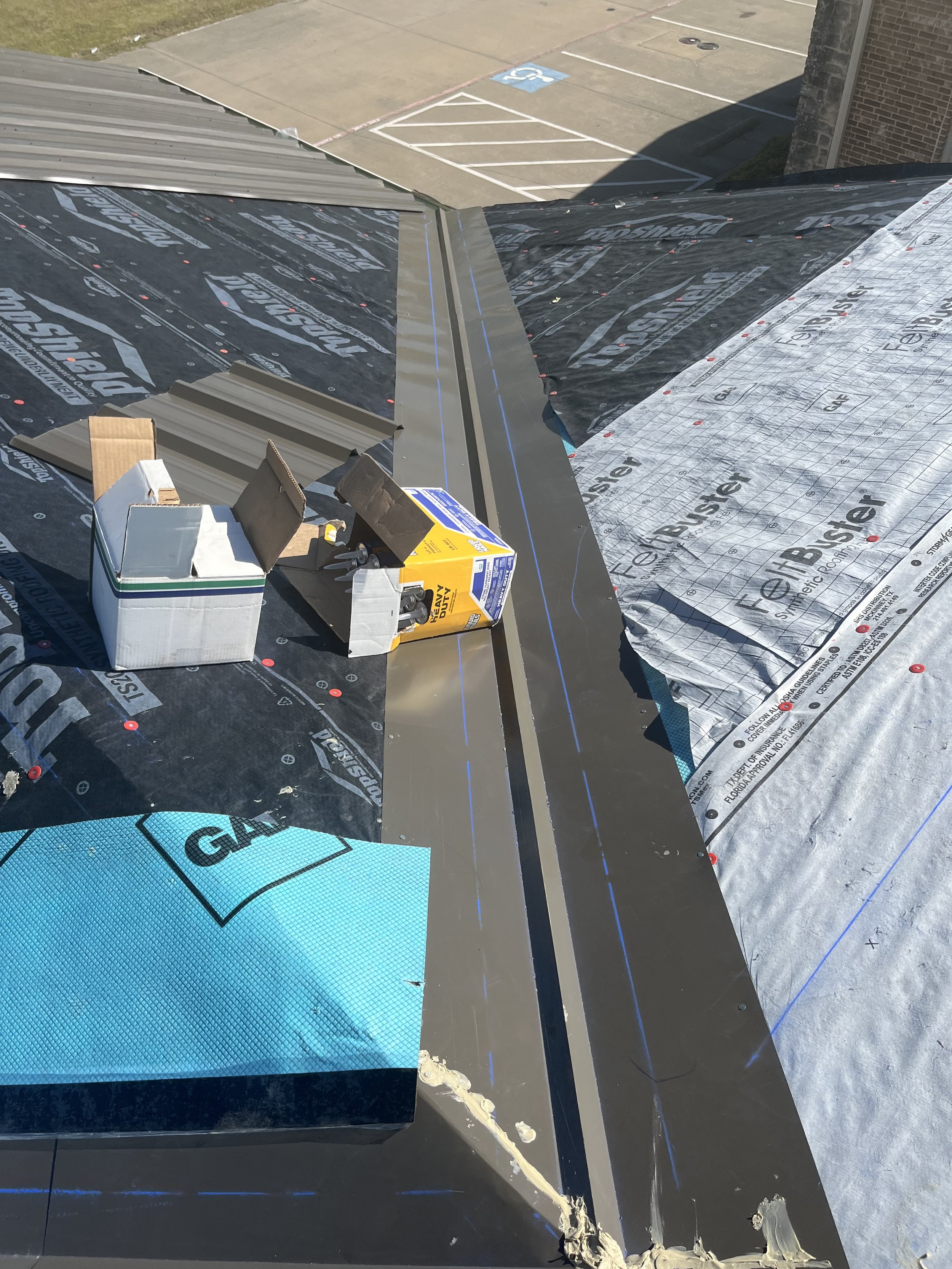 Roof under construction showing roofing materials, tools, and some parking lot in the background.