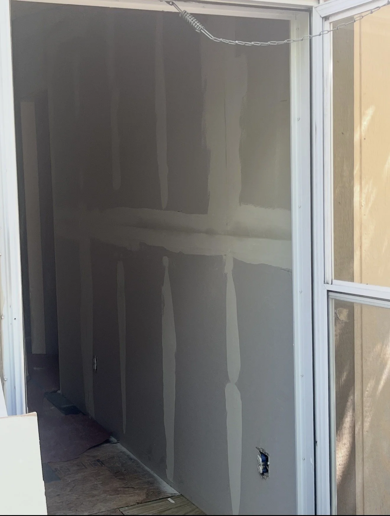 Interior view of a room under construction with drywall installed on the walls and ceiling. An opening with a window frame is visible on the right, and an electrical outlet is seen on the lower right wall. There is construction material on the floor.