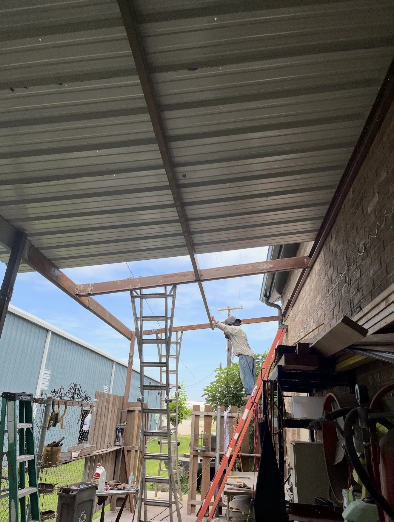 A man working on a construction project under a metal roof extension. He is standing on a ladder, holding a tool, and working on the roof support beams. There are various tools and supplies around, and an outdoor area with a fence and some trees in t
