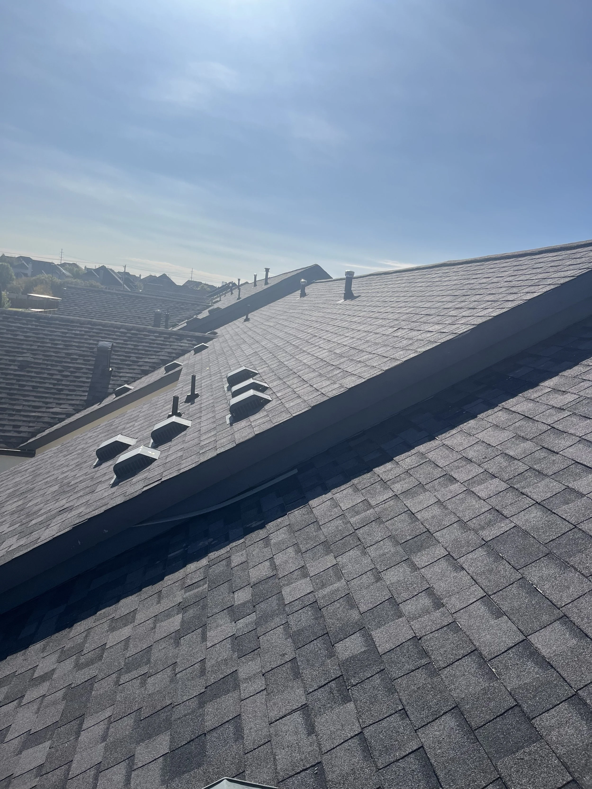 View of rooftops with gray shingles and multiple ventilation pipes under a clear blue sky.