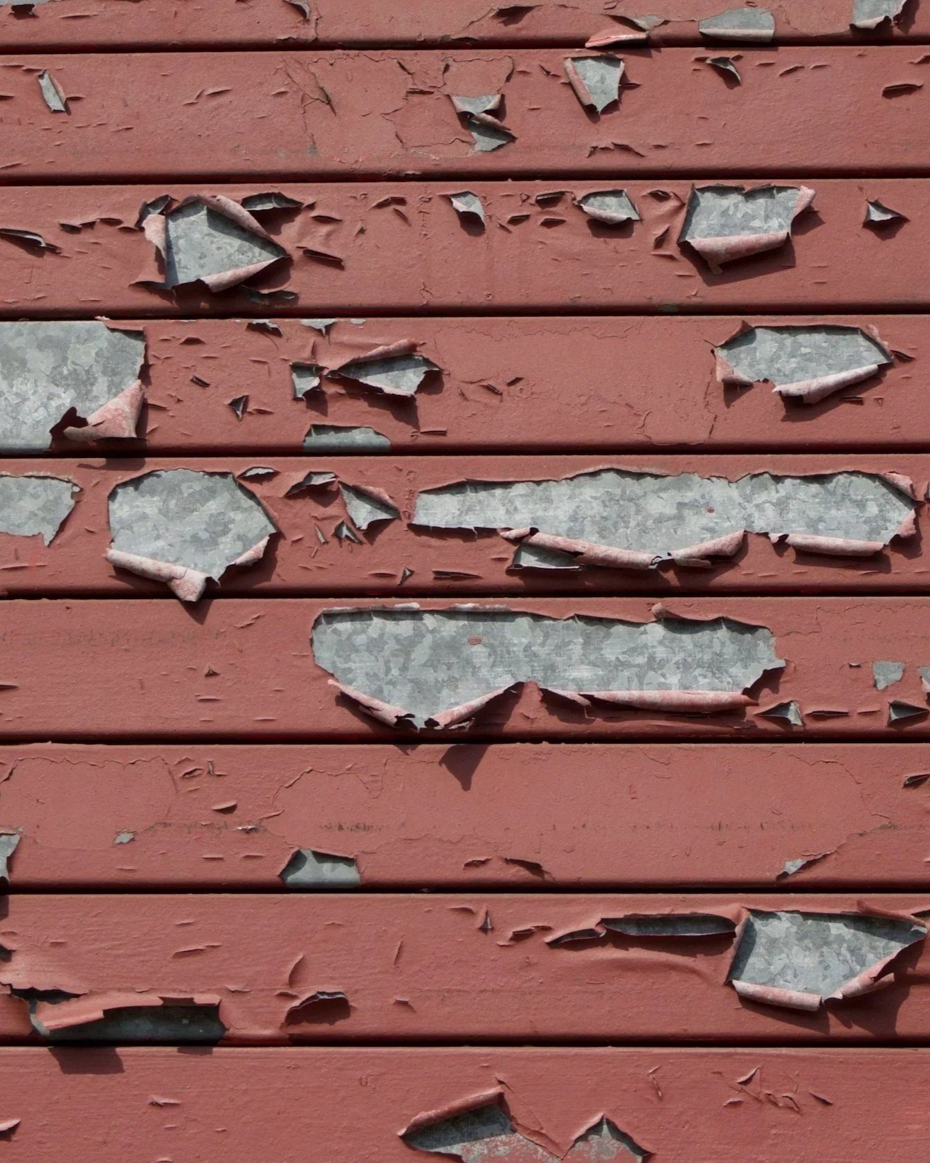 Close-up of a wall with pinkish-red paint peeling off, revealing grayish tiles underneath.