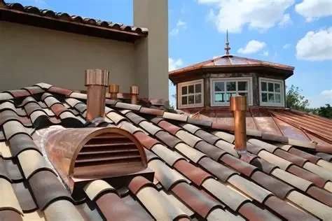 Close-up of a tiled roof with a small vent, chimneys, and a turret in the background under a partly cloudy sky.