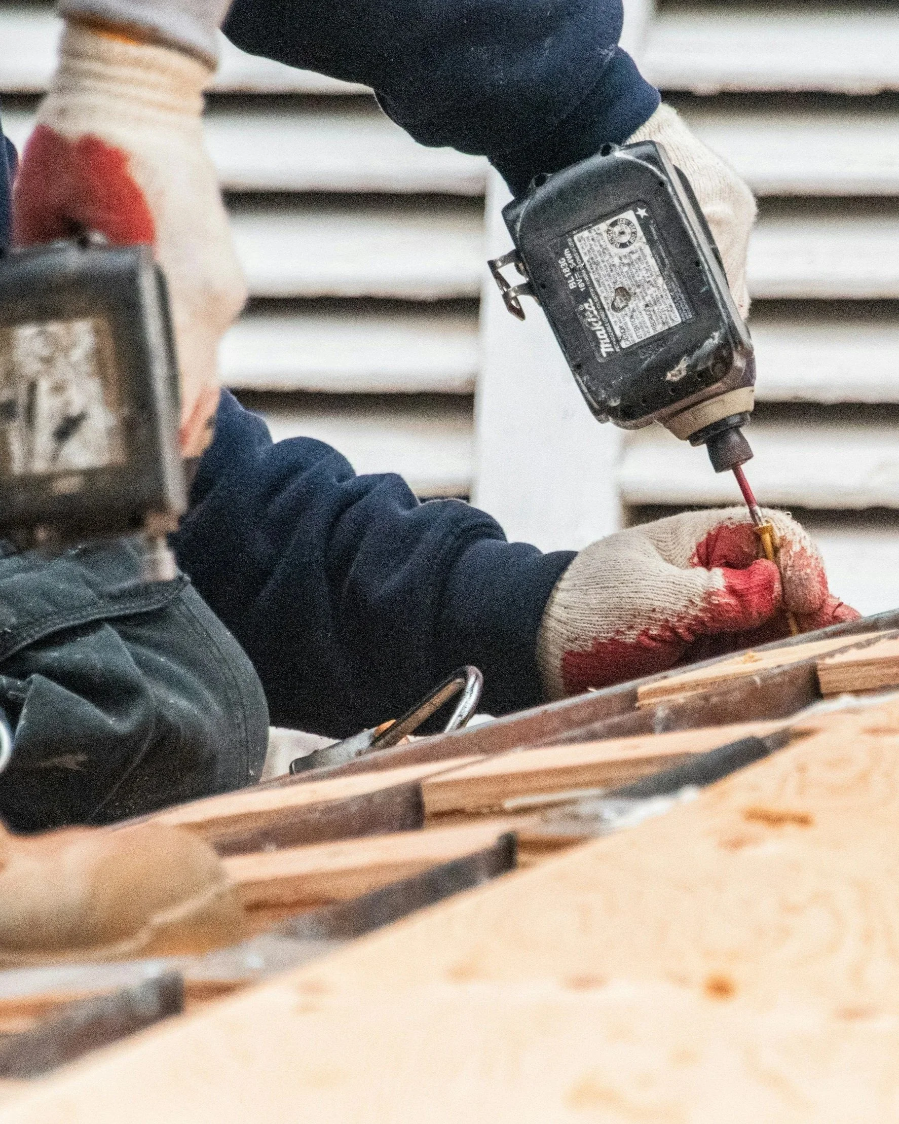 Close-up of a worker's gloved hands using a cordless drill on a piece of wood in a woodworking shop.