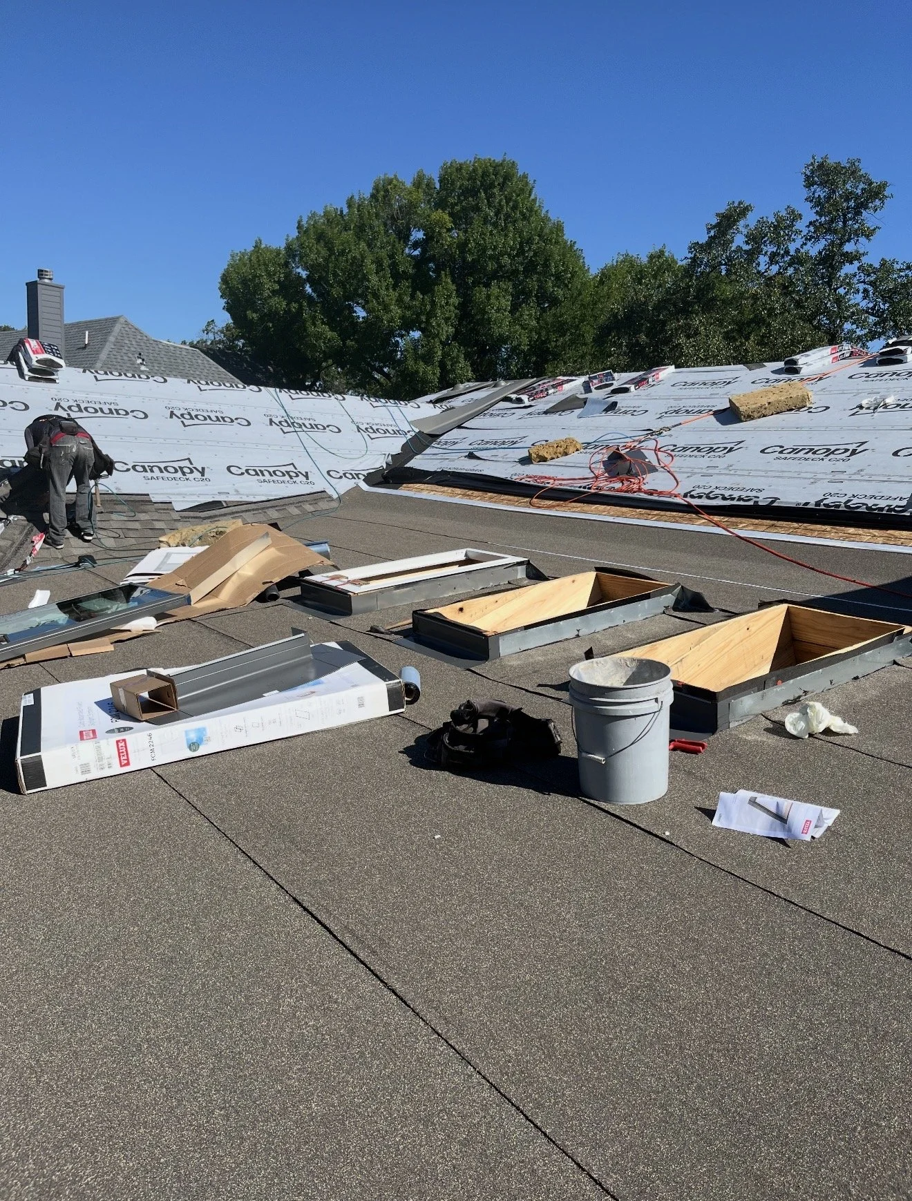 Roof under construction with roofing materials, tools, and supplies scattered on the roof surface. The background features a tree and a clear blue sky.