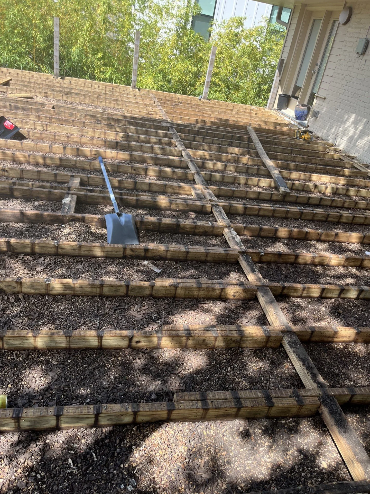 Construction site with wooden framework for a deck or platform, shovels, and gravel, next to a white brick house with a door and windows, and green trees in the background.