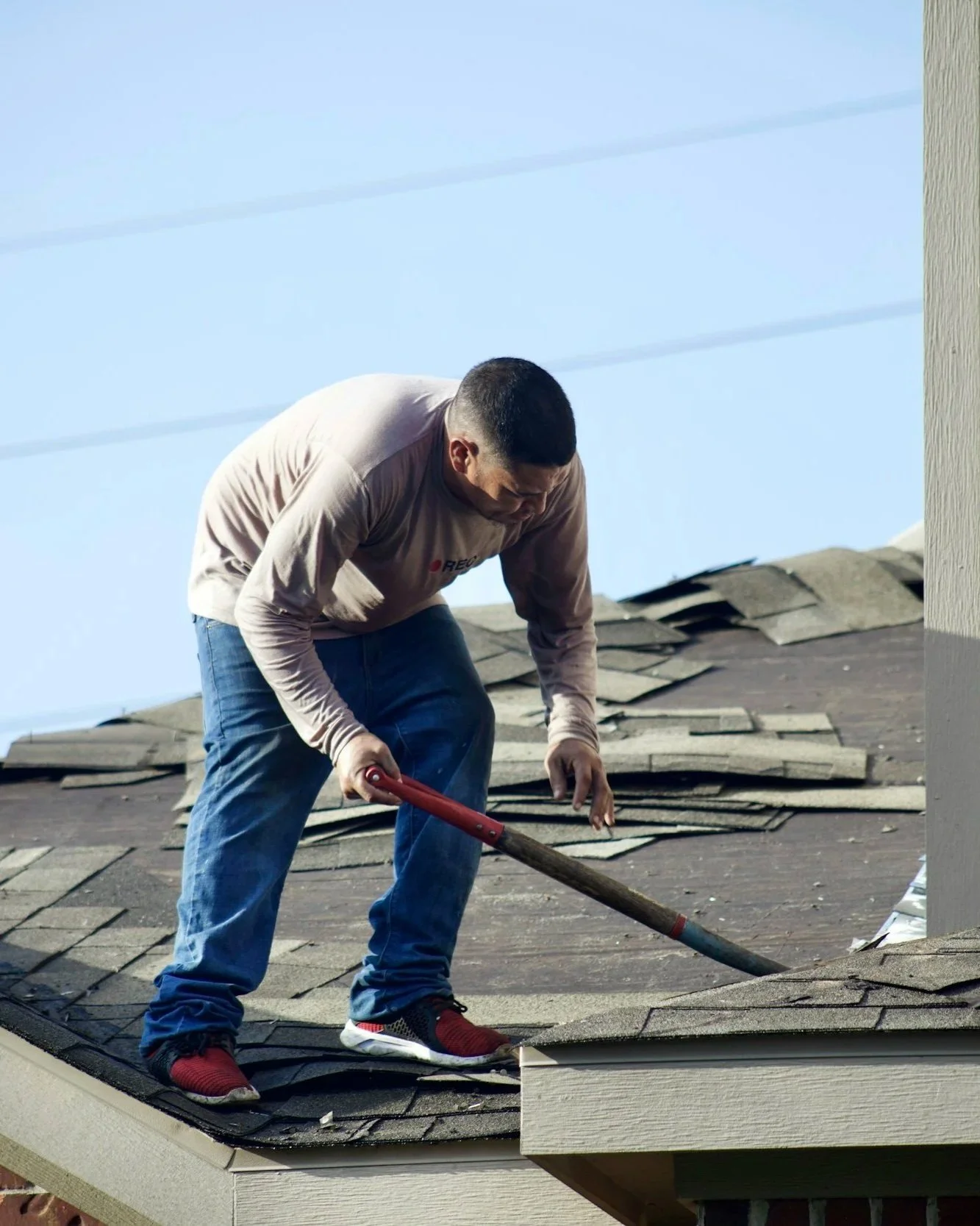 A man working on a house roof, removing old shingles with a crowbar under a clear blue sky.