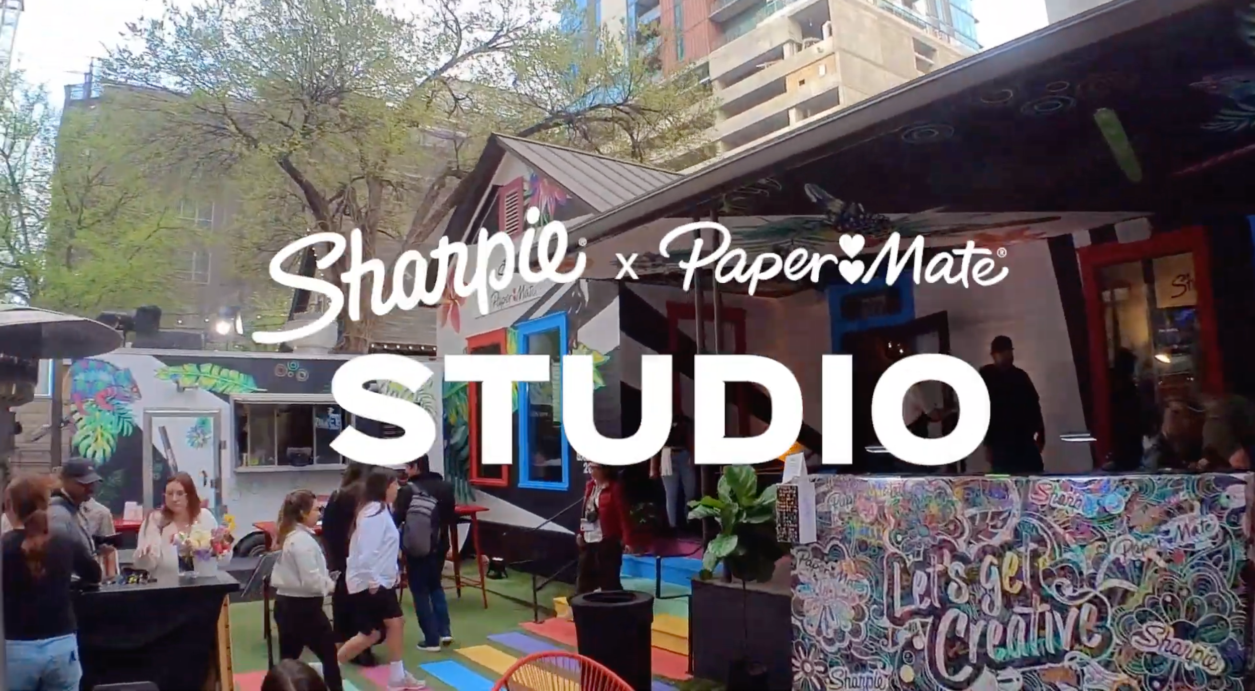 Outdoor scene with a crowd of people at SxSW featuring an art market, booths decorated with colorful artwork, trees with spring leaves, and a sign displaying 'Shapoo x Paper Mate Studio'.