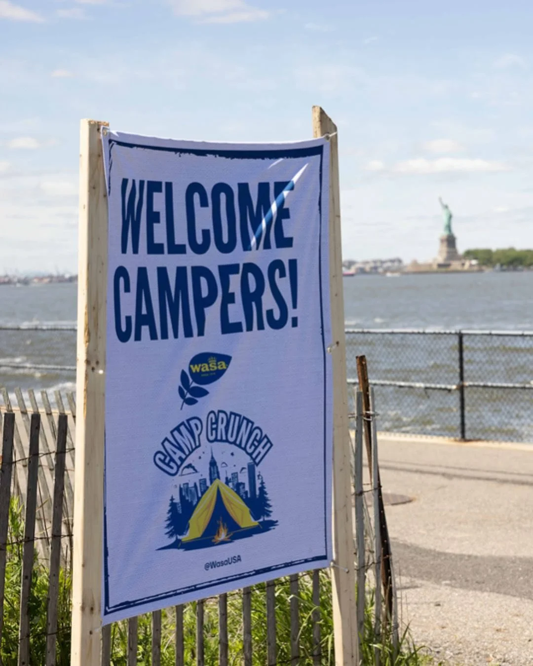 A welcome sign for campers at Camp Crunch by Wasa USA, with the Statue of Liberty visible in the background across the water and a blue sky overhead.