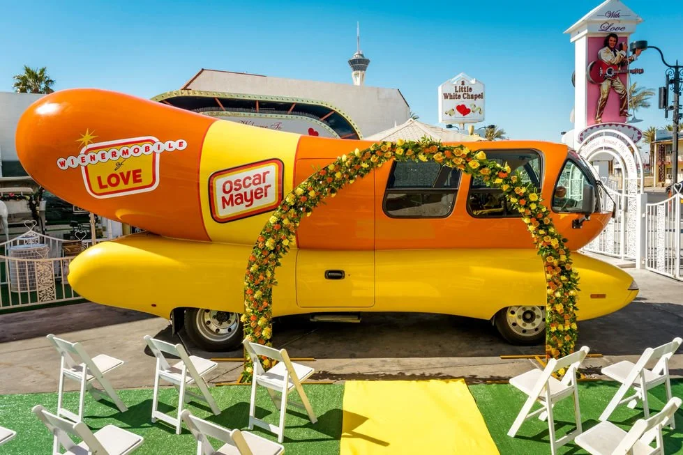A large, colorful Oscar Mayer Wienermobile decorated with a flower arch, set up for a wedding ceremony with white chairs and a yellow aisle in front of a theme park.