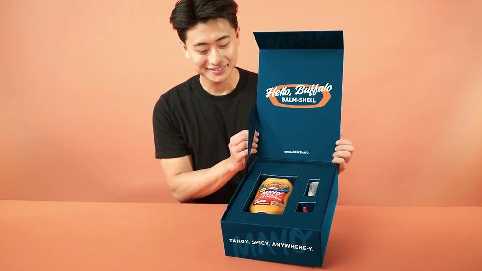 A young man in a black t-shirt opening a blue box that contains a bag of chips, a tube of balm, and a small item, with the box labeled "Hello, Buffalo" and the phrase "Tangy. Spicy. Anywhere-Y." on the front.