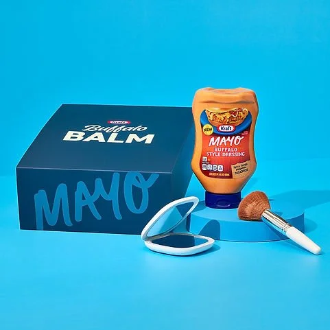 A box labeled 'Buffalo Balm' and a bottle of Kraft Mayo Buffalo Style Dressing, a compact mirror, and a makeup brush on a blue background.