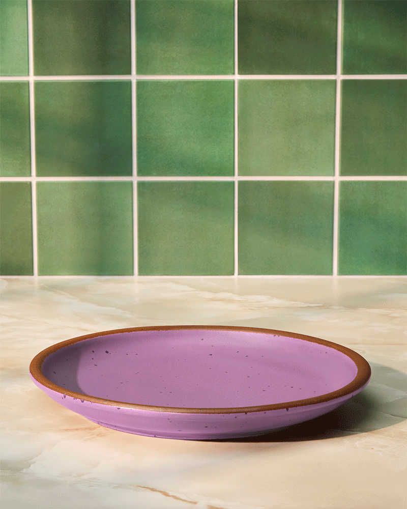 A purple ceramic plate with a brown rim on a light-colored marble countertop against a green tiled wall.