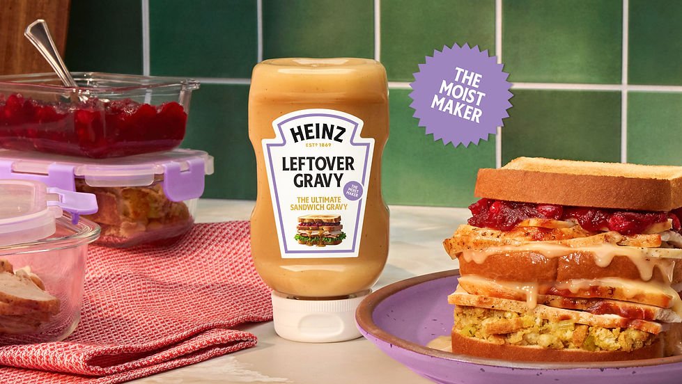 Sandwich layered with various ingredients, a jar of Heinz Leftover Gravy, and containers of cranberry sauce on a countertop with green tiled wall in background.