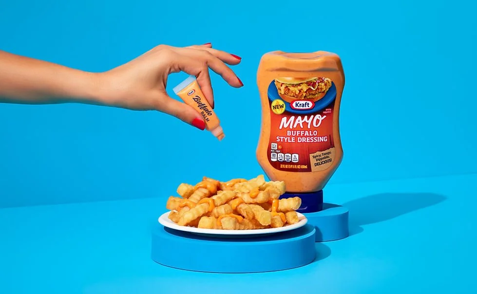 A hand with red nail polish holding a small packet of Buffalo sauce over a plate of crinkle-cut french fries, with a bottle of Kraft Mayo Buffalo Style Dressing in the background on a blue surface.