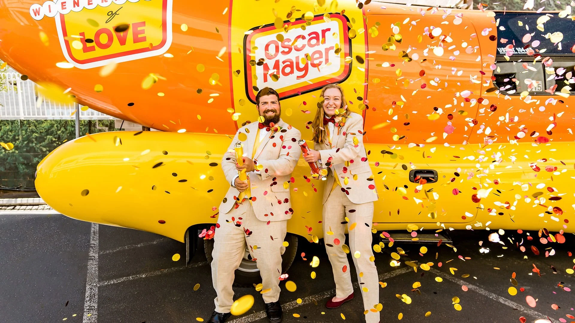 Two Hotdoggers in beige suits standing in front of a yellow bus with the words "Wiener of Love" and "Oscar Mayer" on it, celebrating with confetti and holding hot dogs.