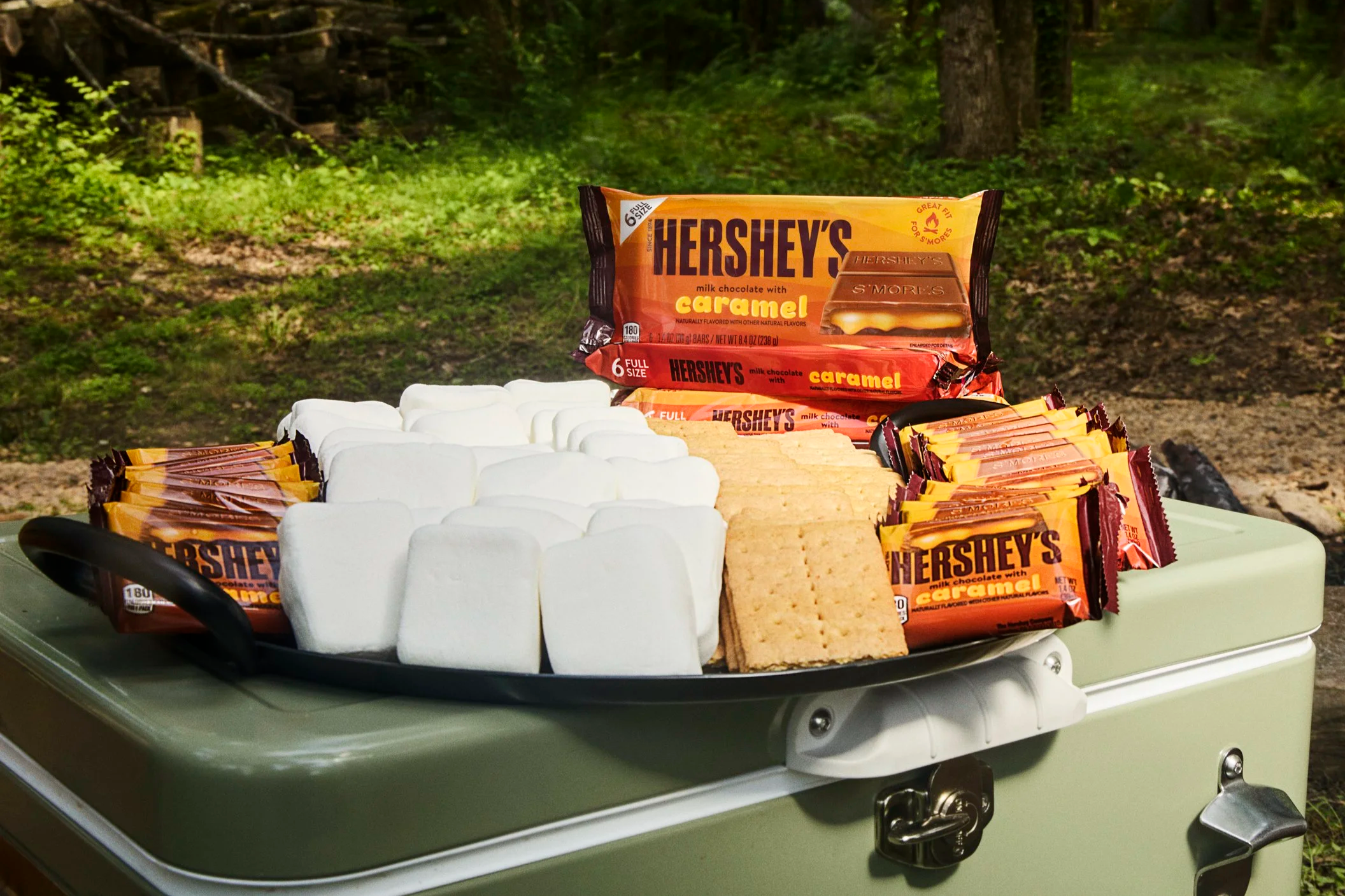 A tray of white marshmallows, graham crackers, and Hershey's milk chocolate bars with caramel on a green outdoor table, surrounded by a wooded area.