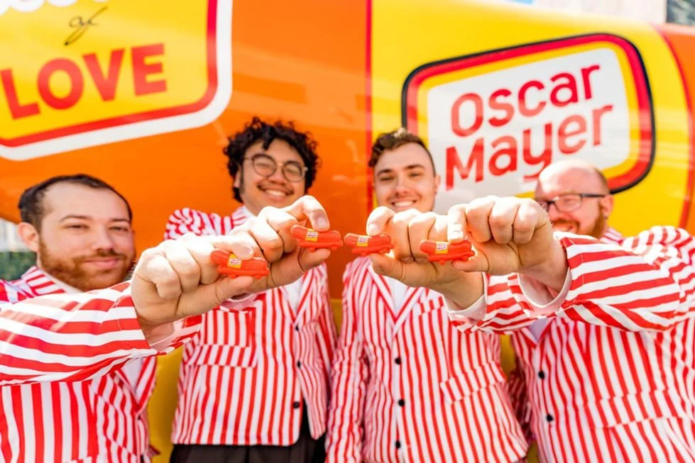 Four men in red and white striped shirts holding orange and red objects, standing in front of a bright orange Oscar Mayer backdrop.