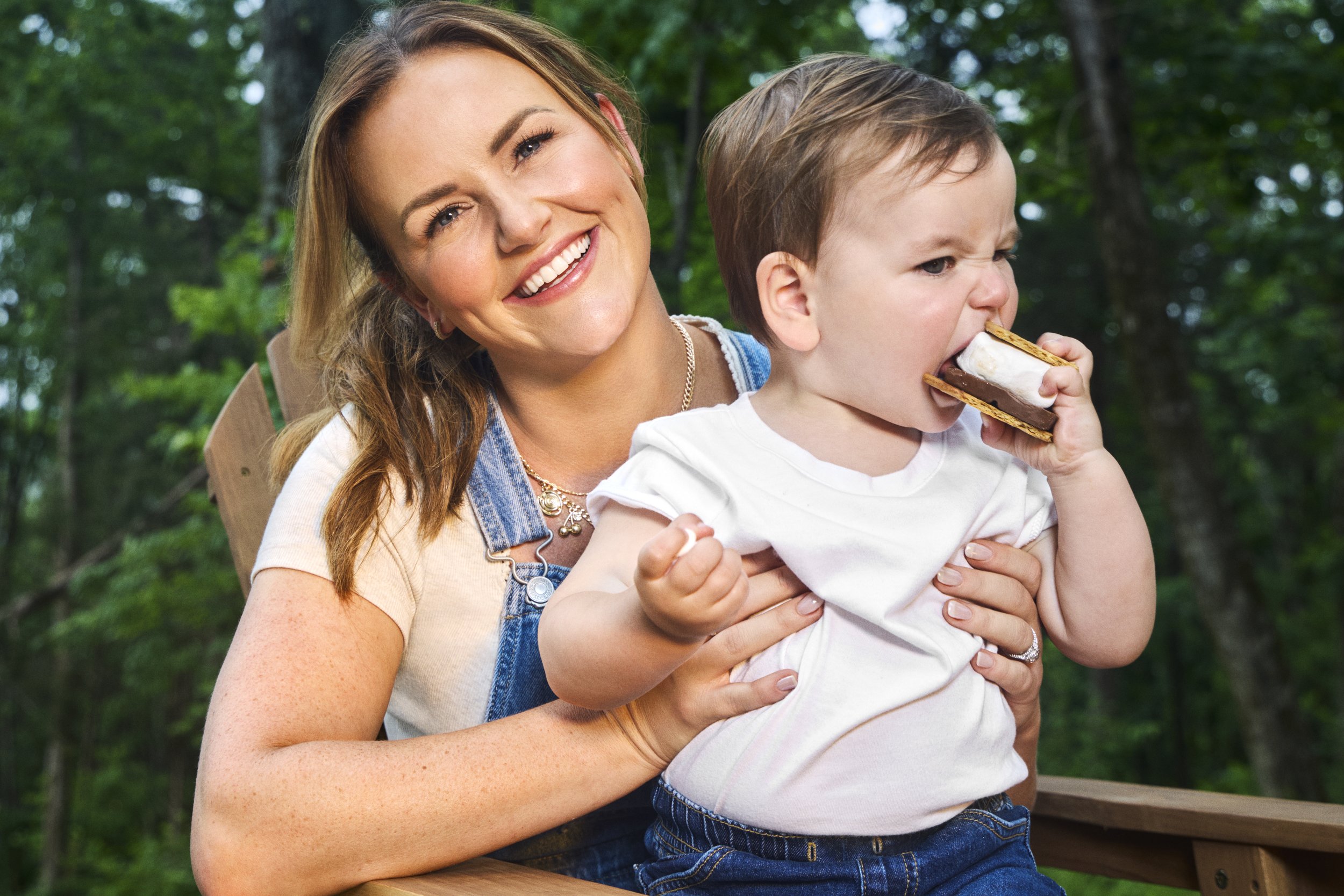 Kailey Dickerson holding a young boy who is eating a s'mores bar outdoors with trees in the background.
