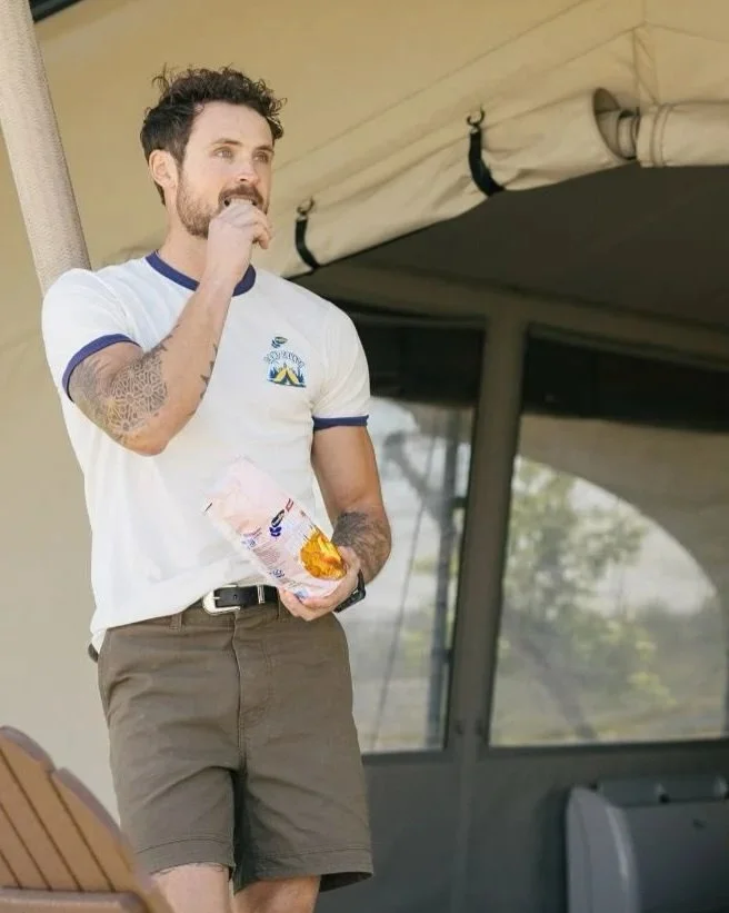 Dylan Efron standing outside, holding a snack bag in one hand and eating with the other. He is wearing a white T-shirt with blue trim and tan shorts.